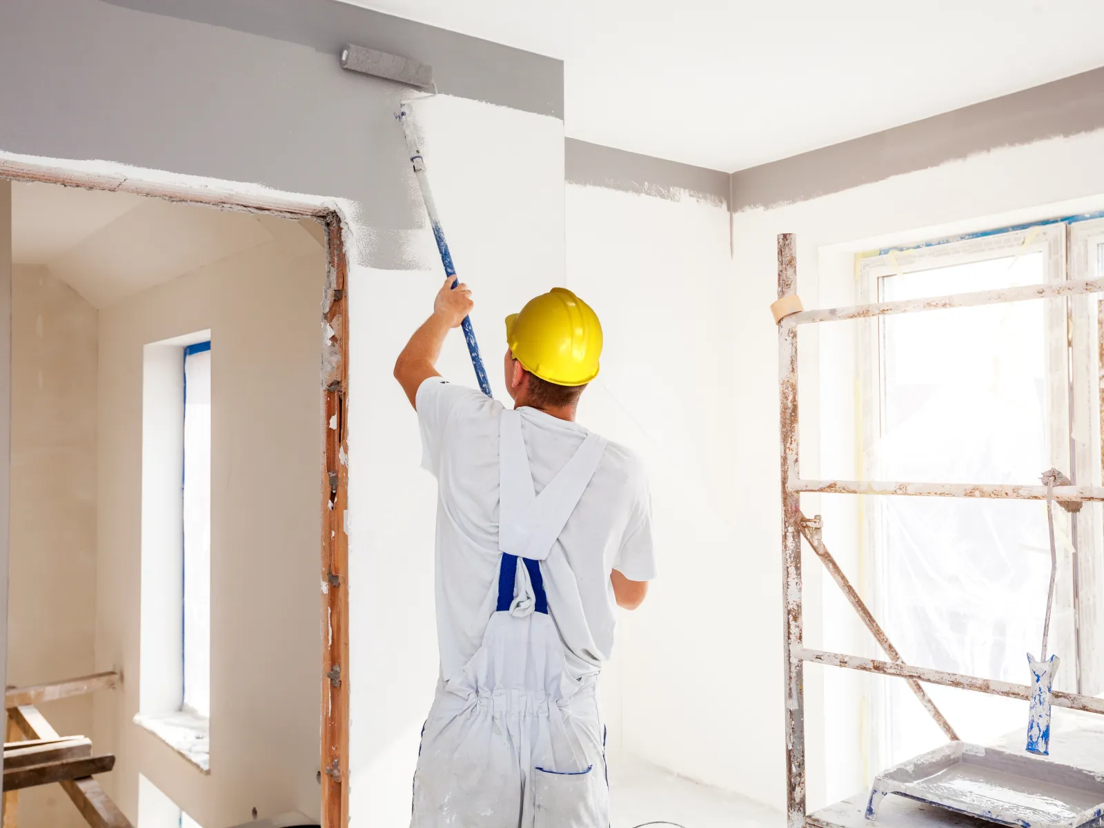 Construction worker in white overalls and yellow helmet painting interior wall with roller in bright room.