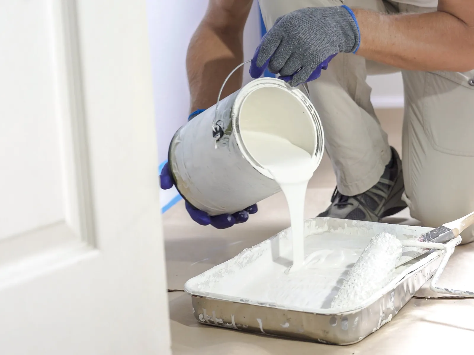 Person pouring white paint from a can into a tray with a paint roller nearby on protected floor.