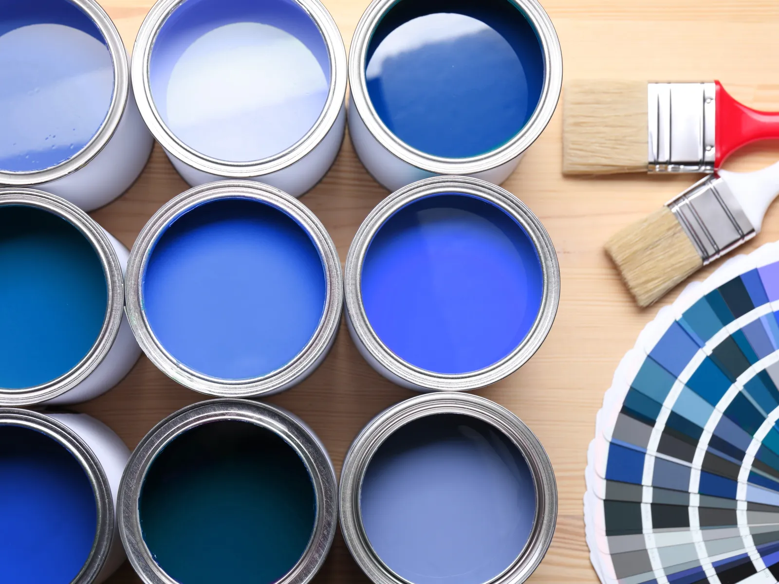 Open cans of blue paint with brushes and a color swatch fan on a wooden surface showcasing blue shades.