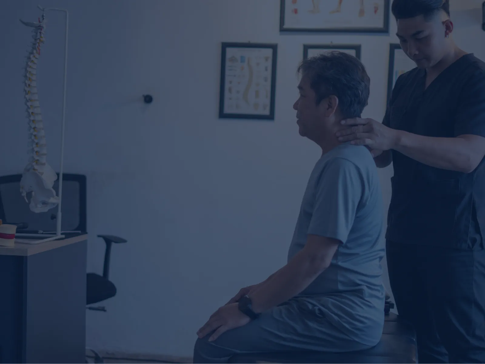 Chiropractor performing neck adjustment on seated middle-aged man in clinic with spine model and anatomical charts.