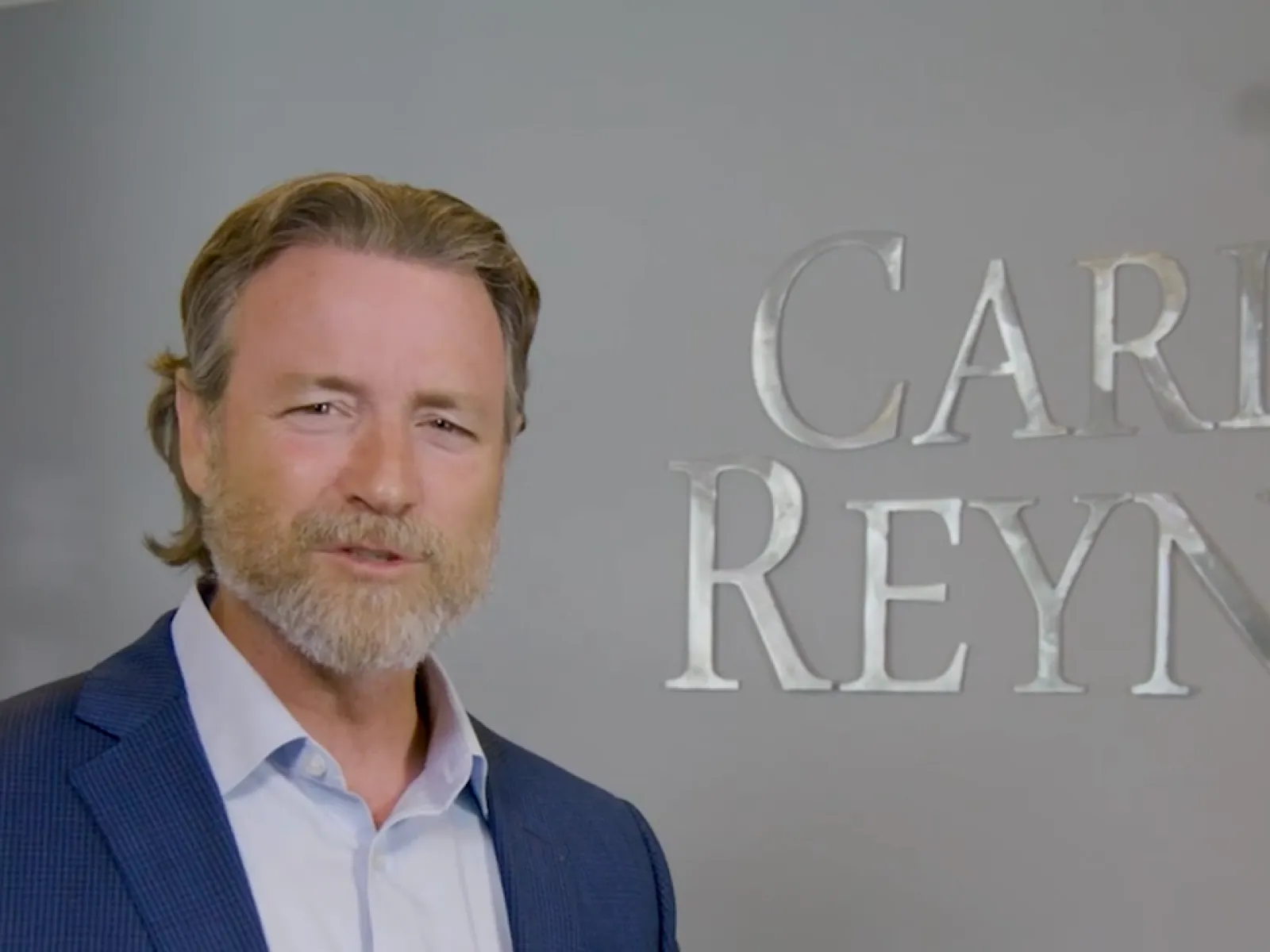 Man in blue blazer stands in front of a gray wall with partially visible silver letters in an office setting.