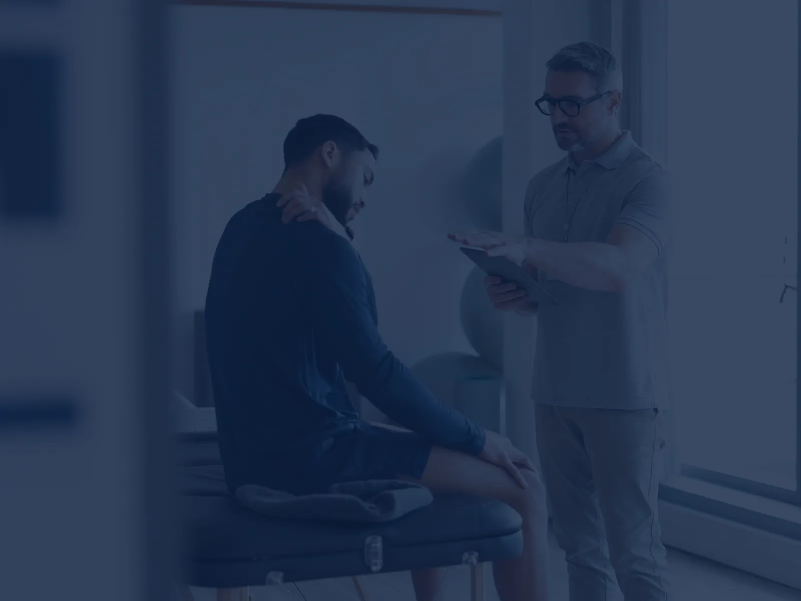 Physical therapist consulting with male patient sitting on treatment table in bright clinic room