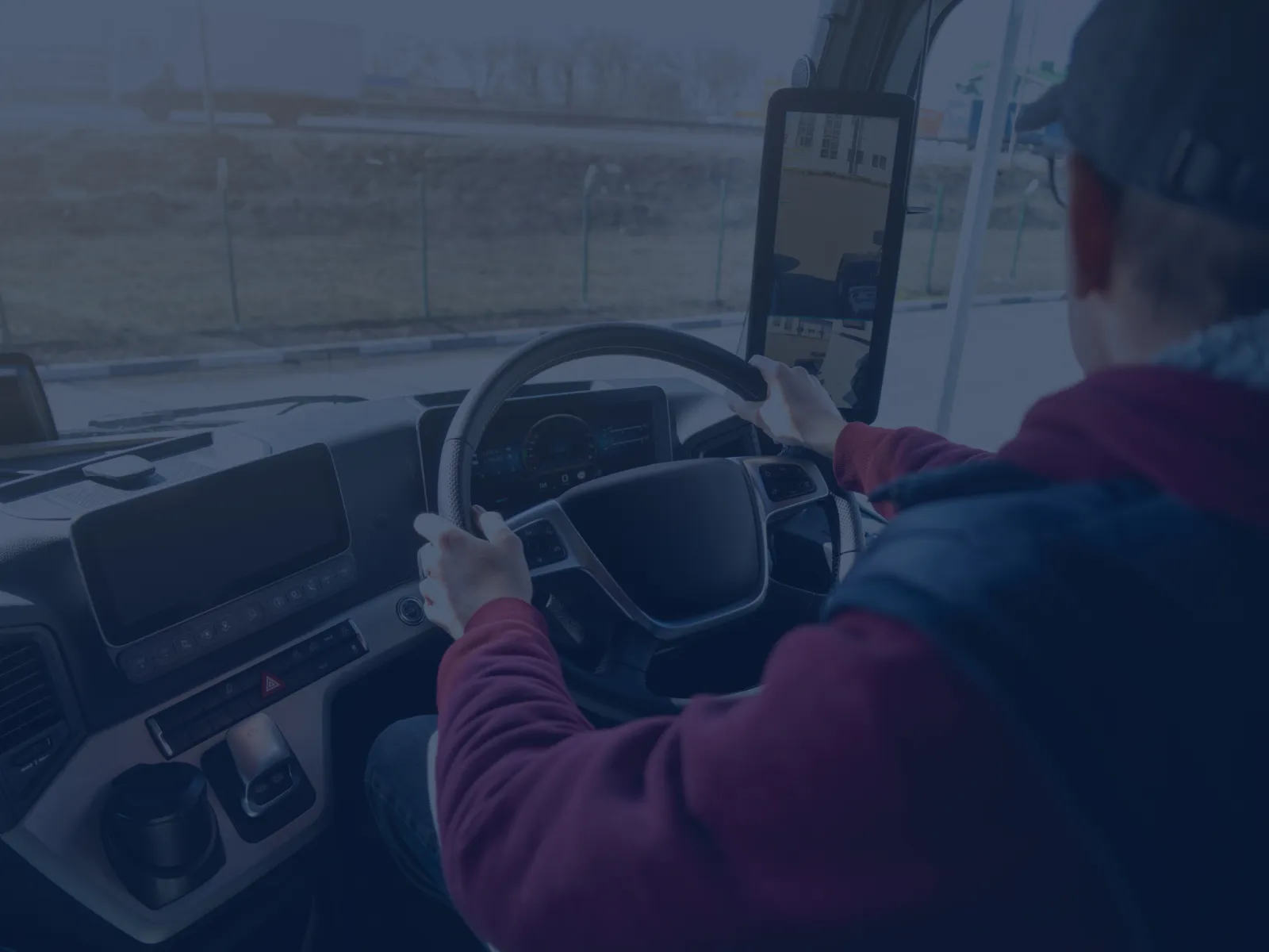 Driver wearing cap and jacket operating steering wheel of a modern truck interior during daytime.
