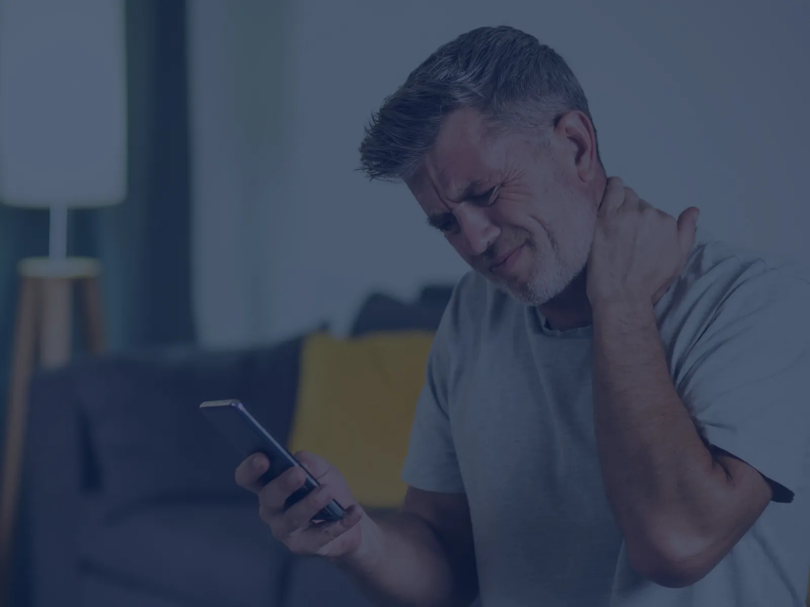 Middle-aged man in gray shirt holding phone and rubbing neck, sitting on couch in dimly lit room.