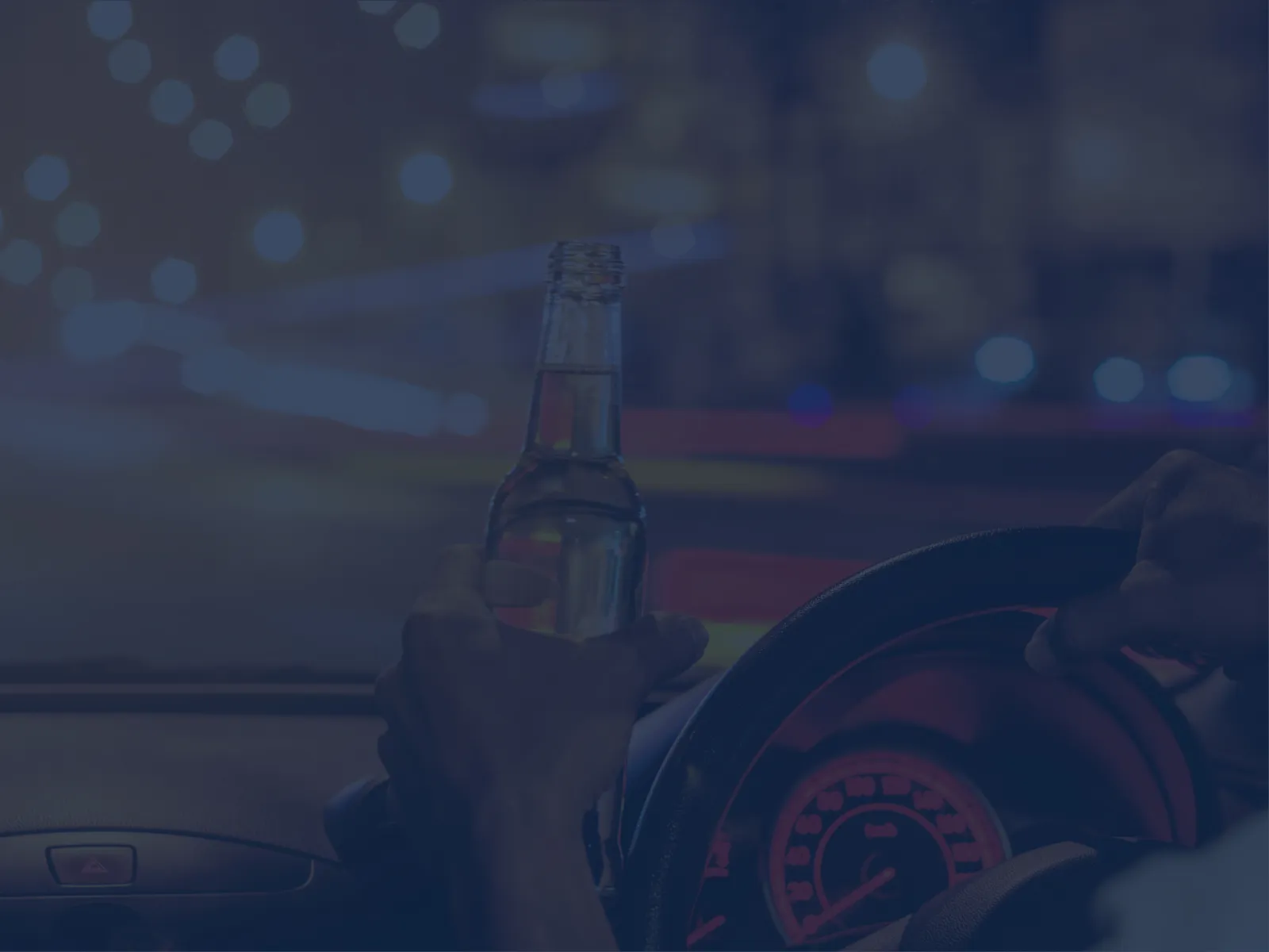 Person holding a beer bottle while driving a car at night with blurred city lights on the road.