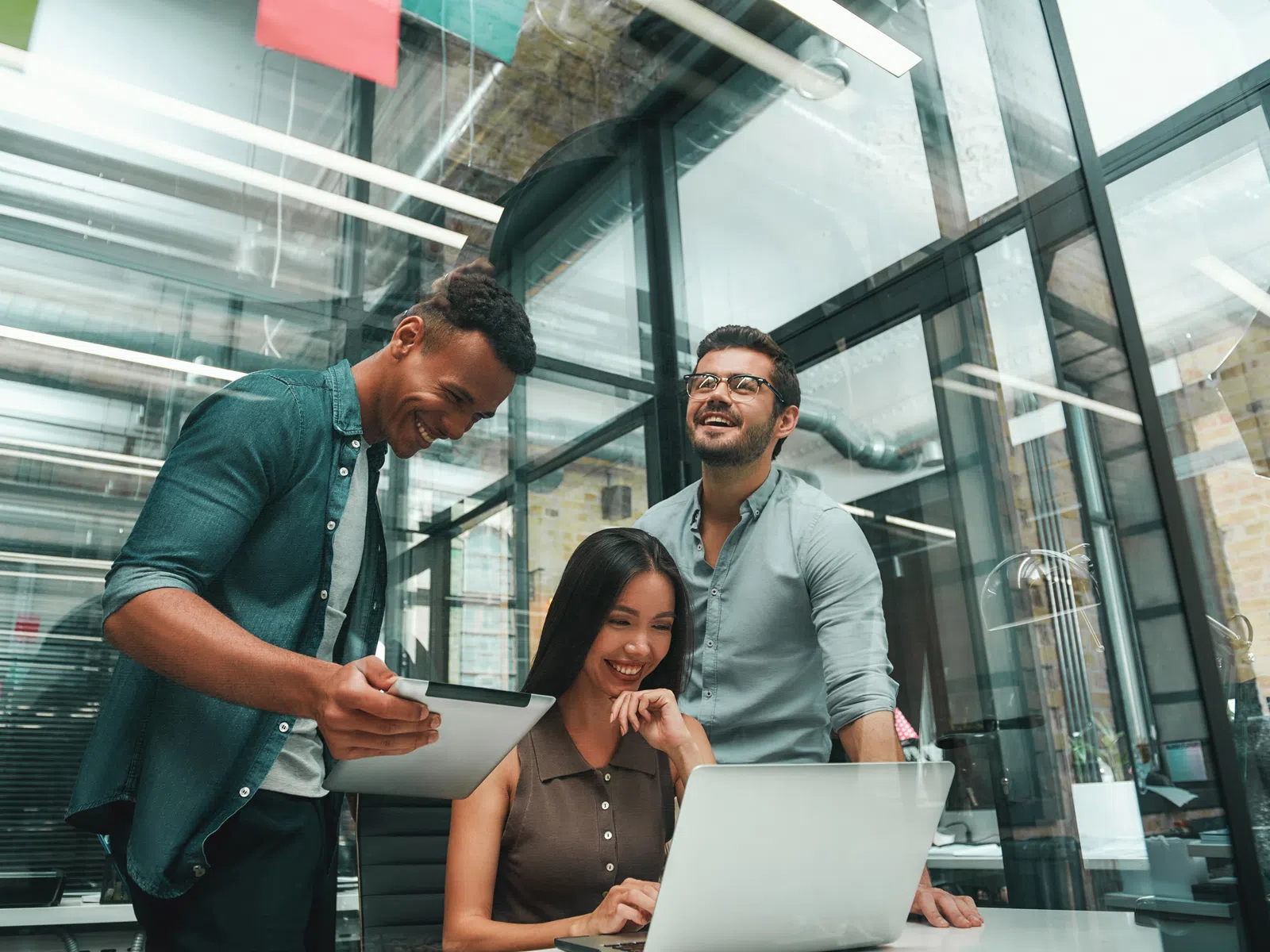 Three coworkers smiling and collaborating in a modern glass office with a laptop and a tablet.