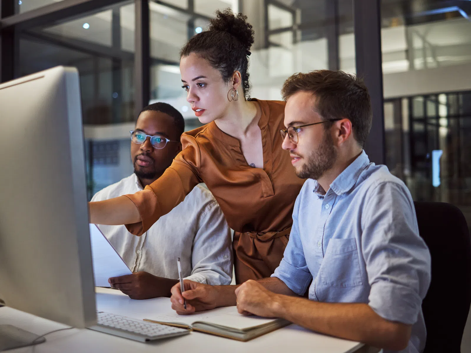 Three diverse coworkers collaborate over a desktop computer in a modern office during evening hours.