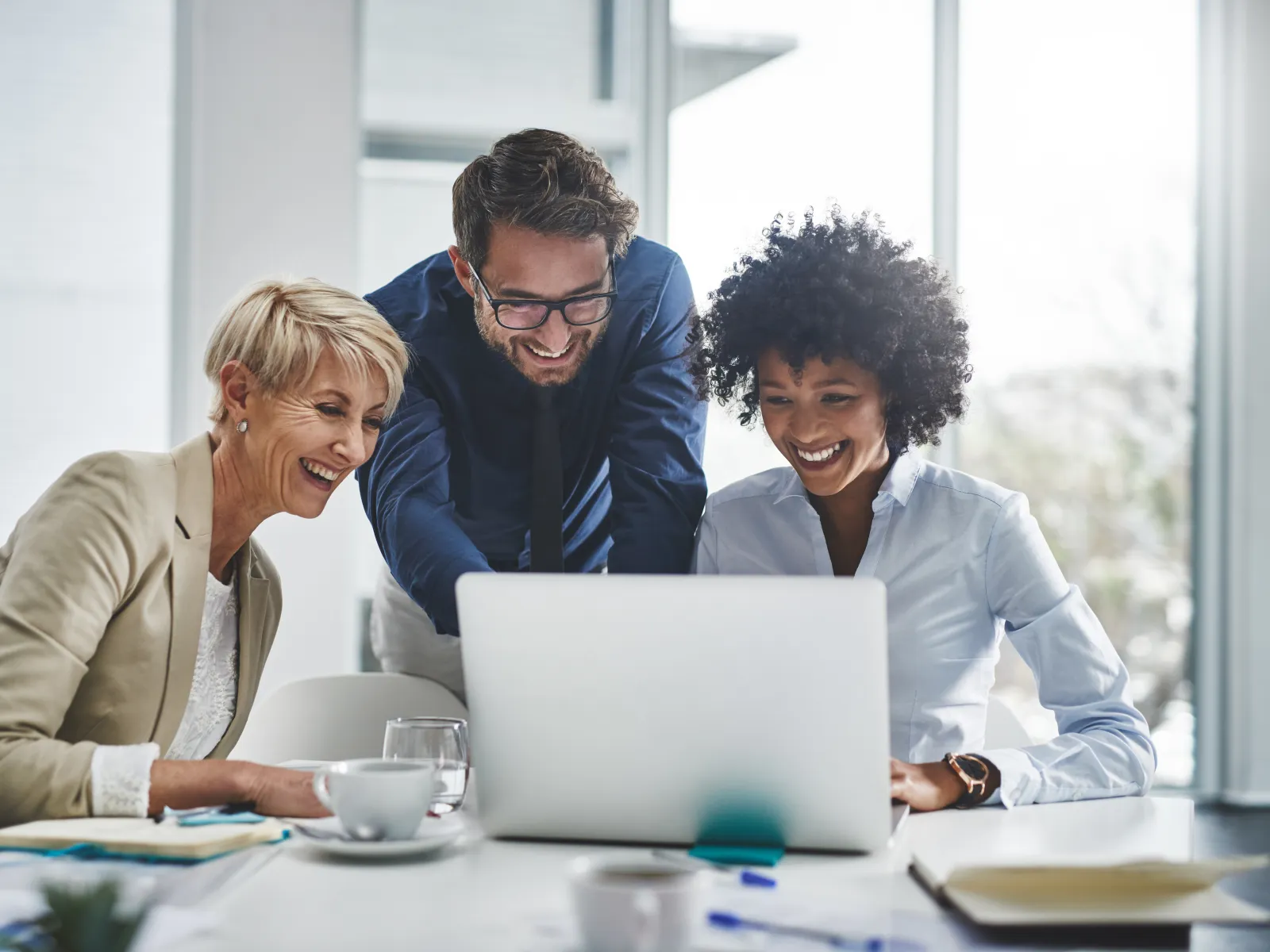 Three diverse professionals smiling and collaborating over a laptop in a bright modern office meeting room.