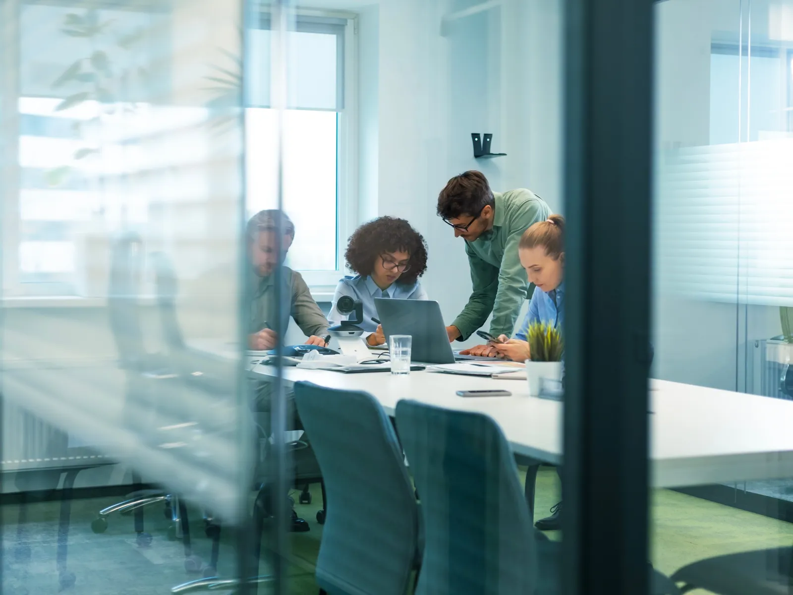 Team of four professionals collaborating around a laptop in a modern office meeting room with glass walls.