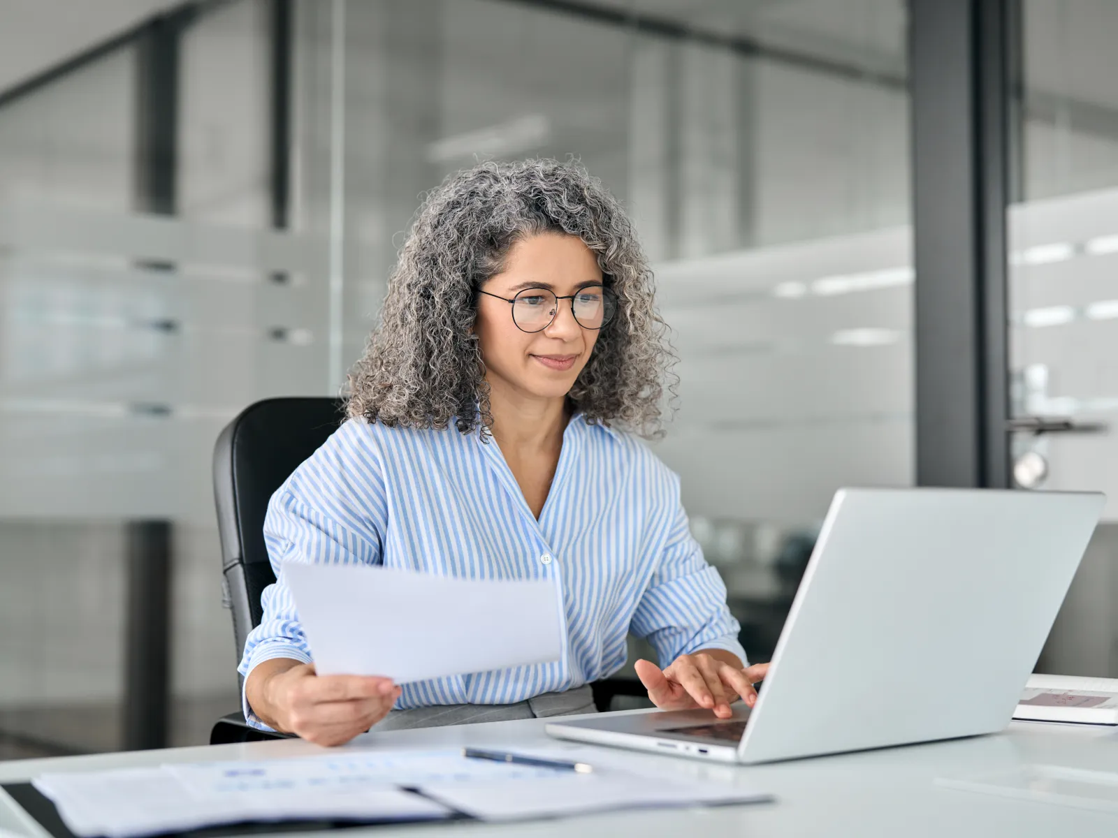 Smiling woman in glasses working on laptop and holding document in modern office setting.
