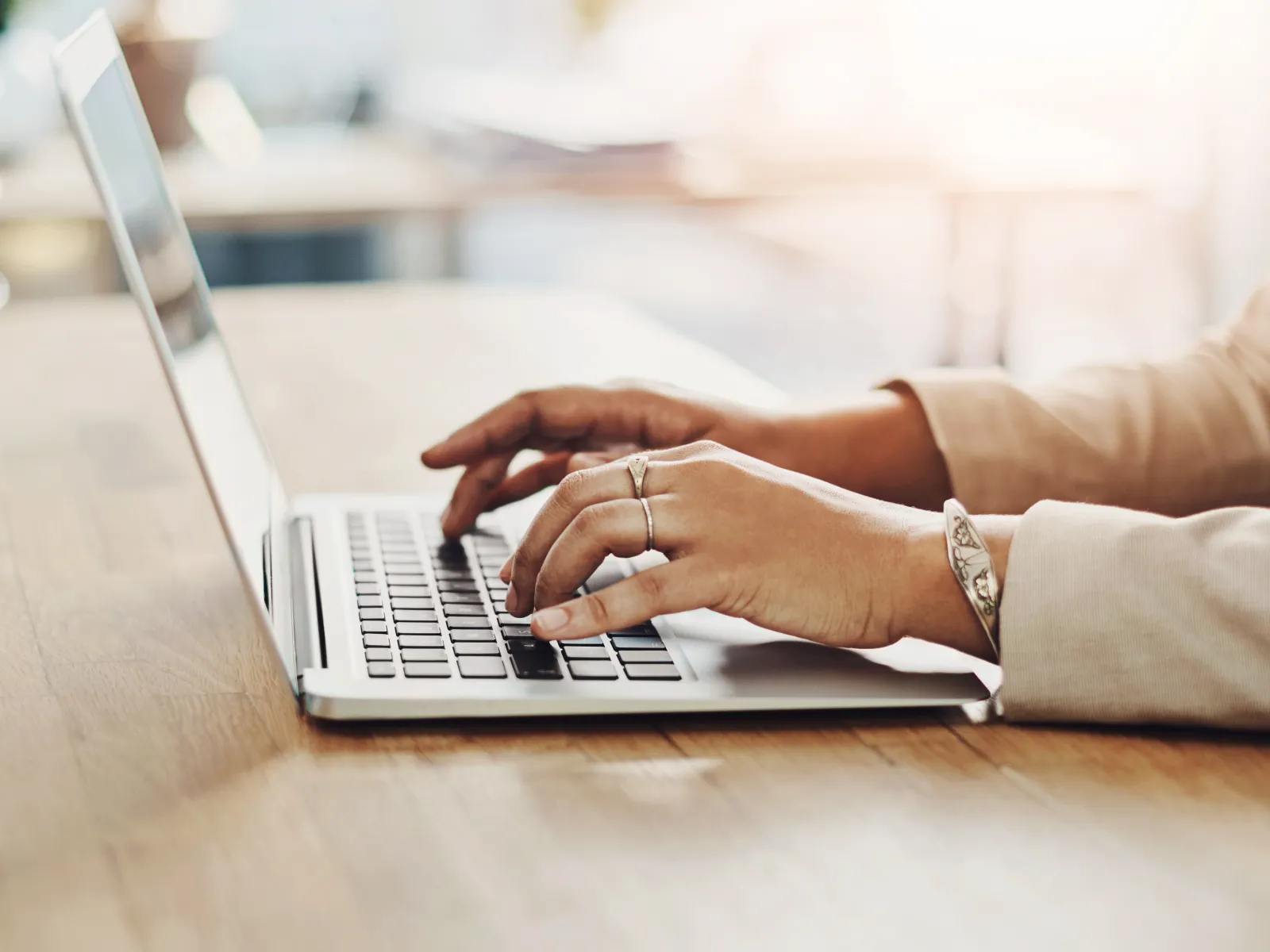 Person typing on a laptop keyboard with natural light and wooden desk in a bright office space