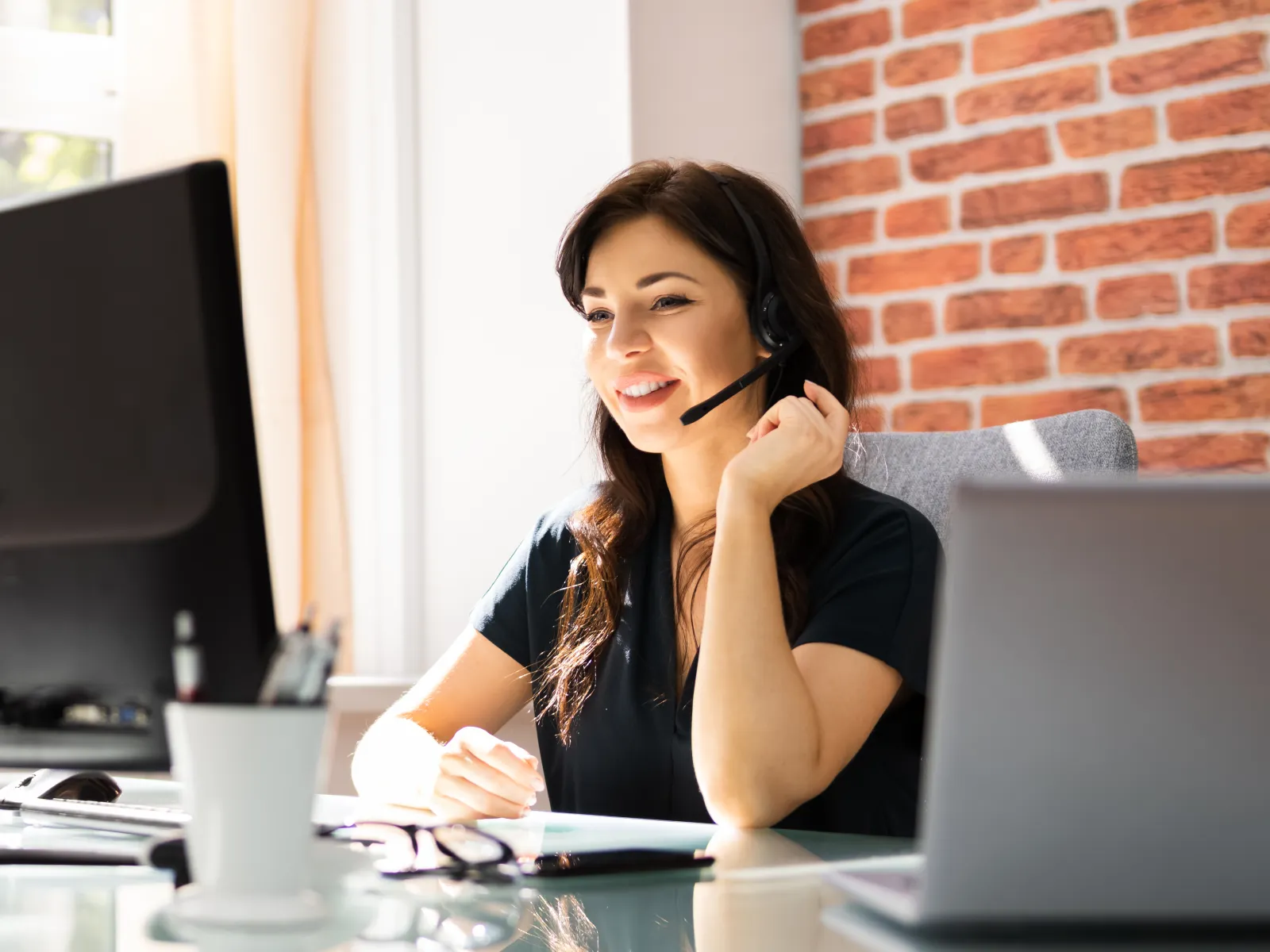 Woman wearing headset smiling while working on computer at bright office desk near window