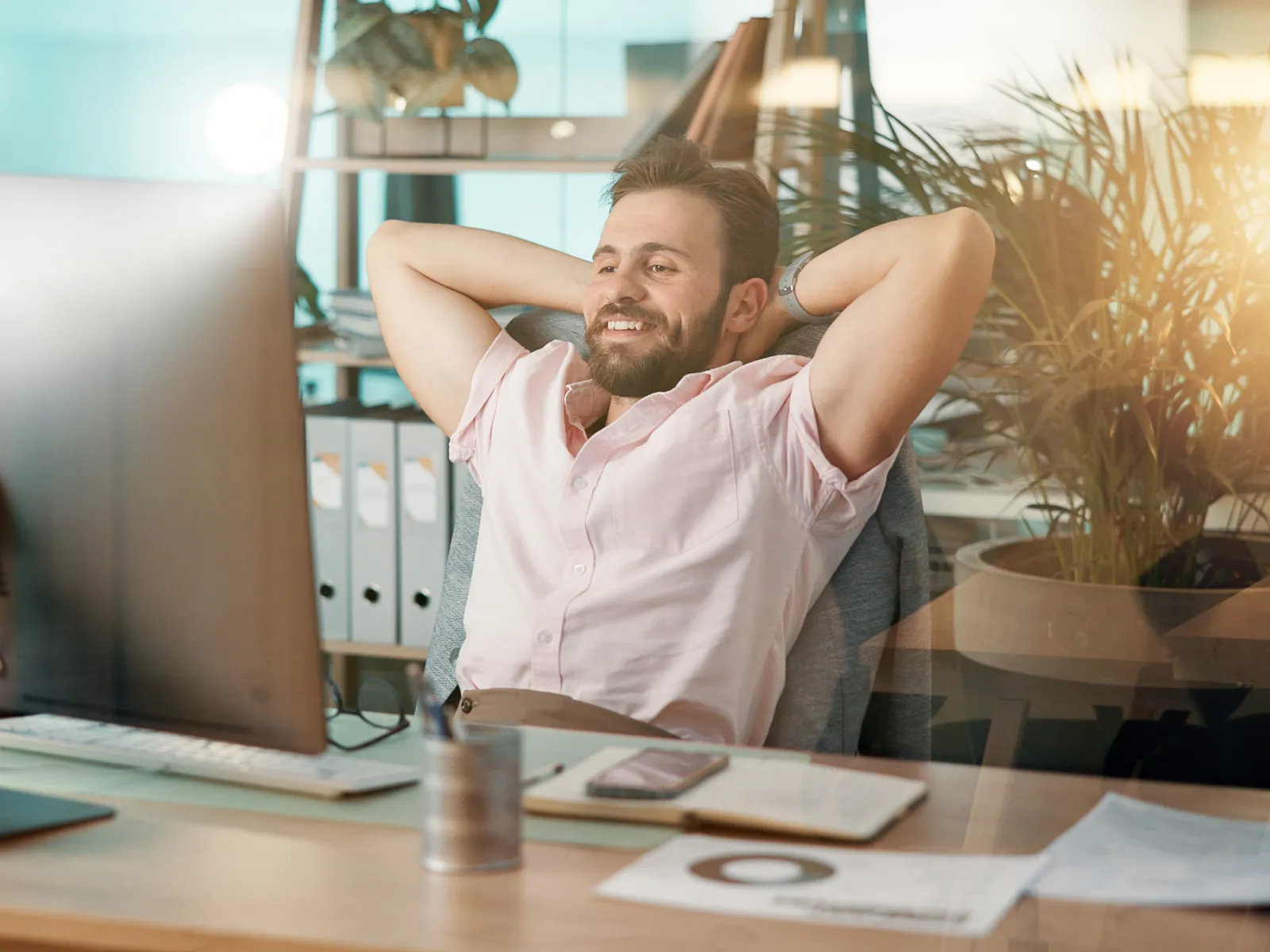 Smiling man relaxing at desk with computer in modern office, arms behind head, sunlight and plants in background