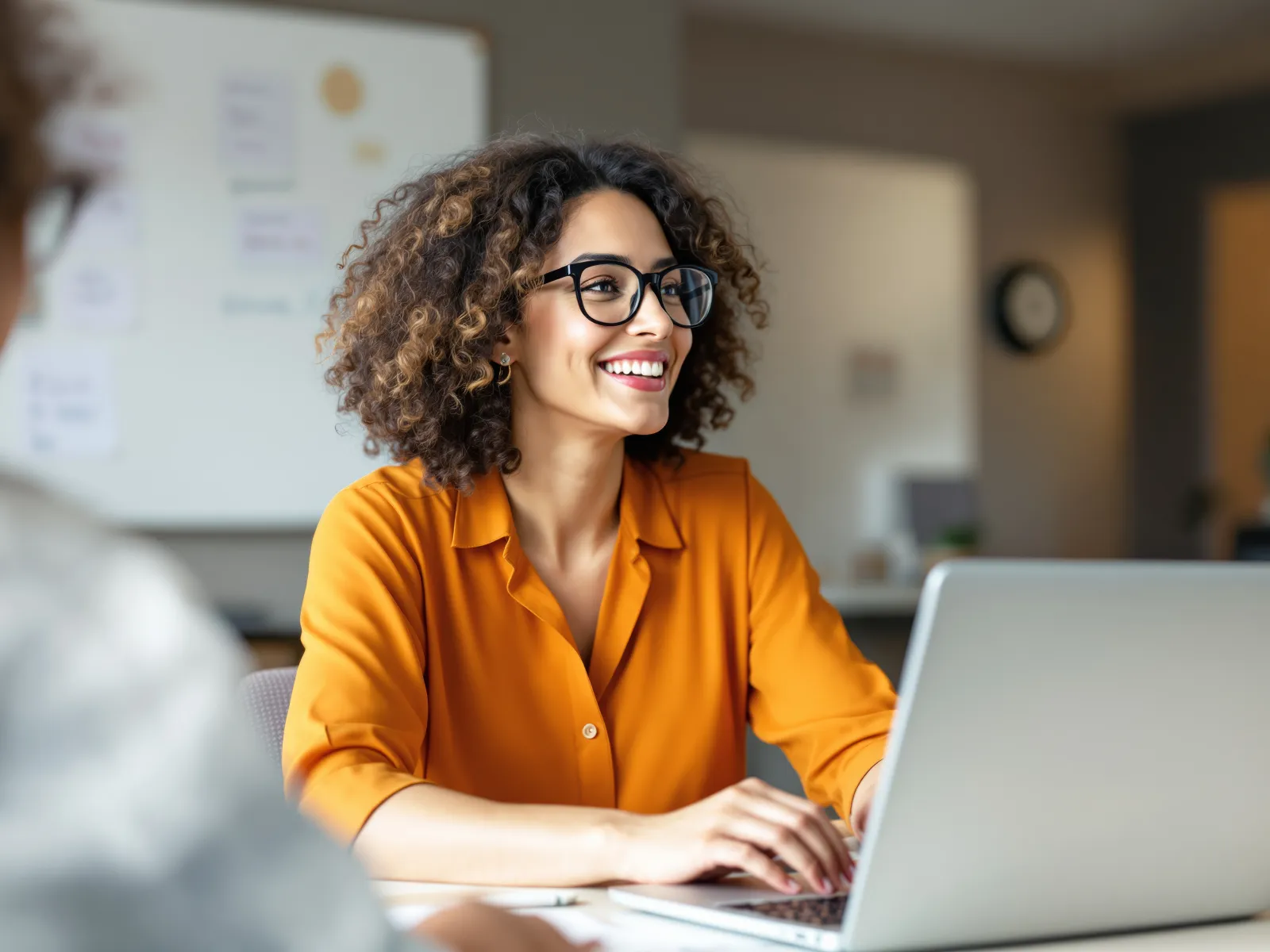 Smiling woman with glasses in orange shirt working on laptop in modern office setting.