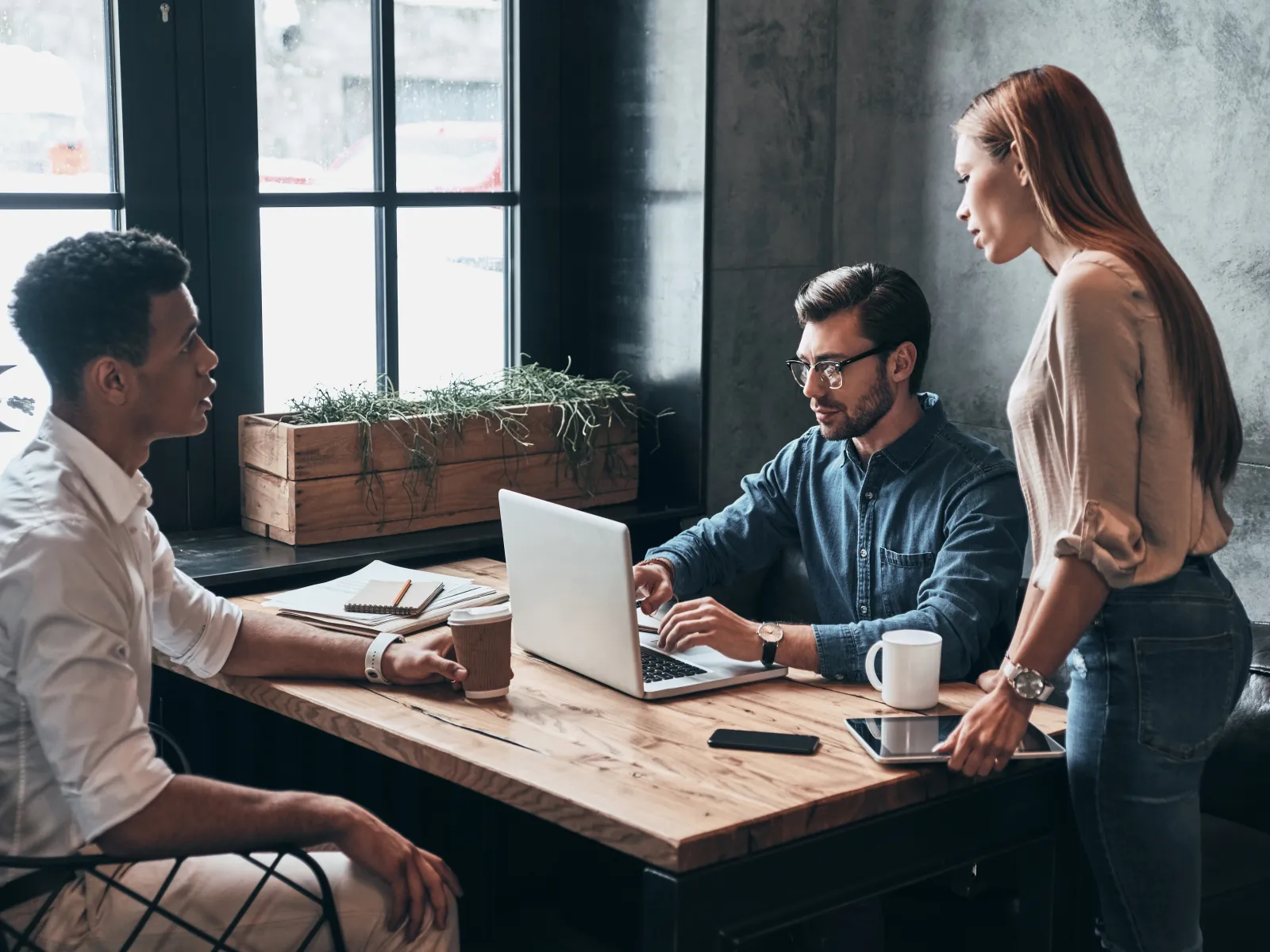 Three young professionals collaborating at a wooden table with laptop and coffee in a modern office setting