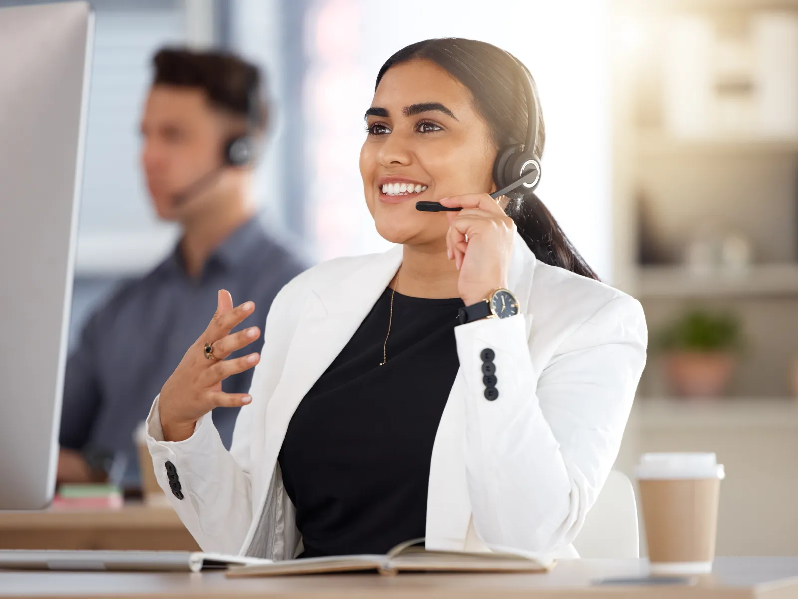 Smiling customer service representative wearing headset and white blazer working at desk with computer and coffee.