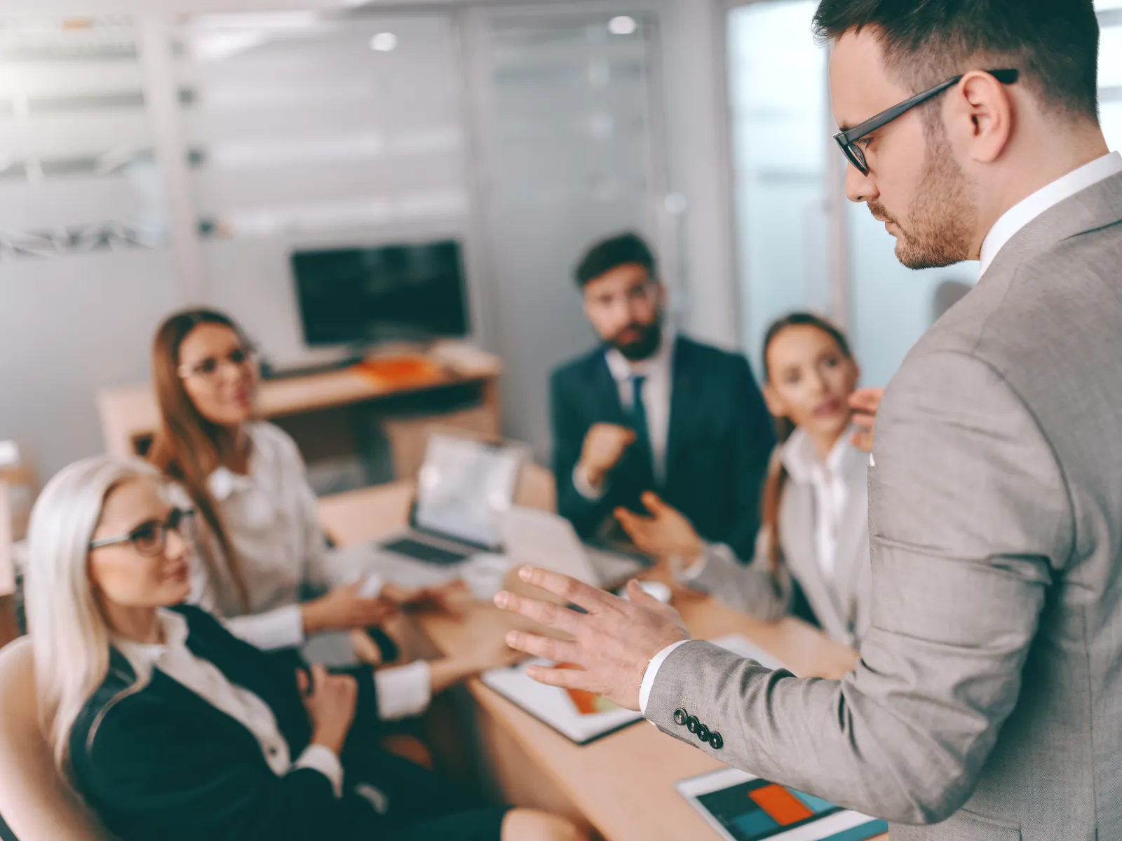 Businessman in gray suit presenting to a group of colleagues in a modern office meeting room.