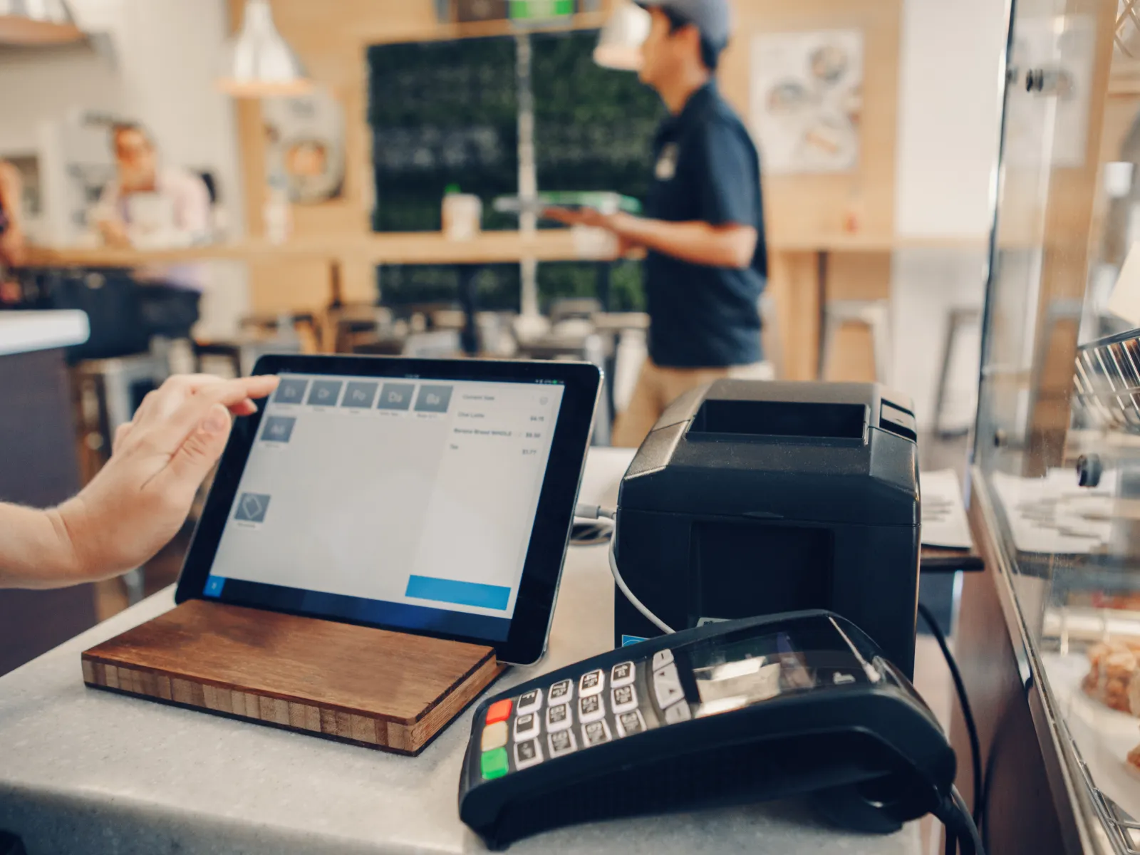 Hand interacting with a touchscreen POS system in a cafe with card reader and printer on counter