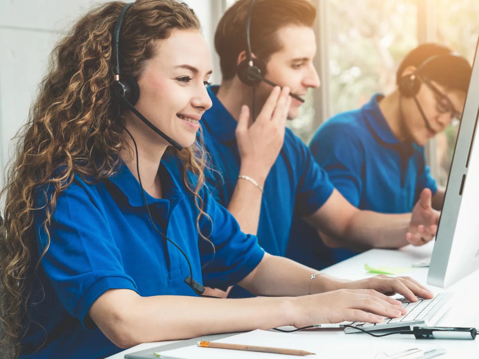 Customer service team in blue uniforms wearing headsets working at computers in a bright office.