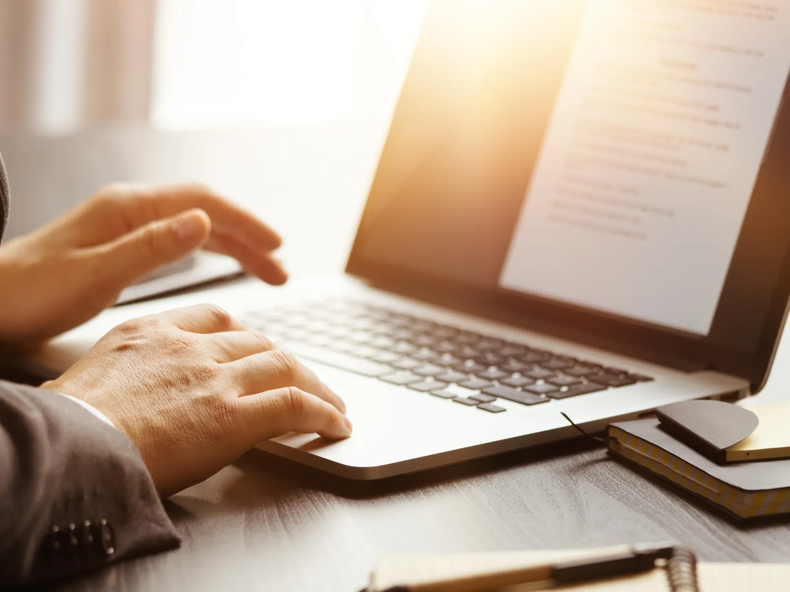 Person in business attire typing on laptop with document on screen and notebook with sticky notes nearby