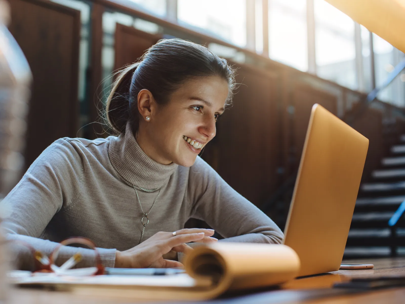 Smiling woman in gray turtleneck working on laptop in bright room with natural light and staircase background
