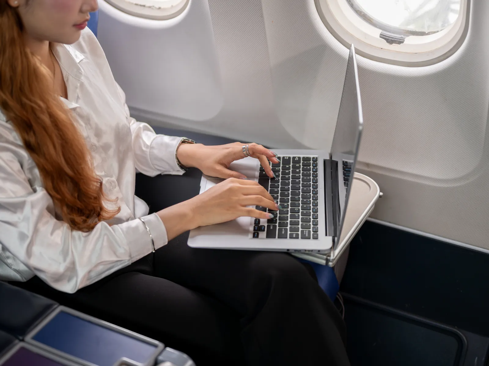 Woman working on a laptop while seated by the window inside an airplane cabin during a flight.
