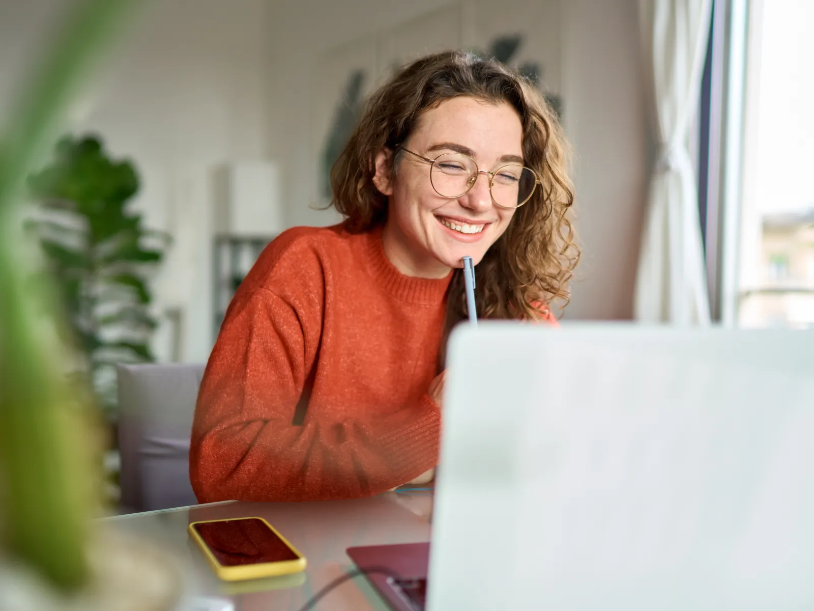 Smiling woman in glasses and orange sweater working on laptop in bright cozy room with plants and natural light.
