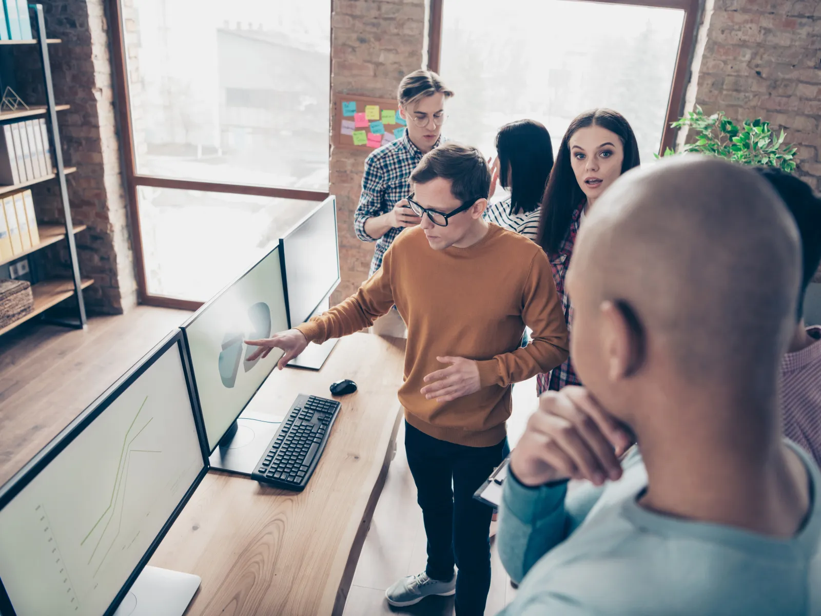 Young professionals discussing data charts on computer screens in modern office with brick walls and large windows