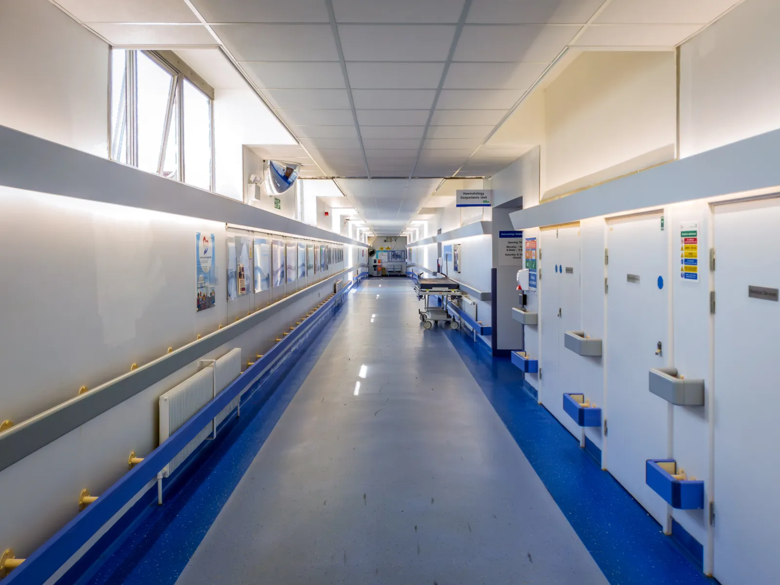 Empty hospital corridor with blue flooring, white walls, medical equipment, and closed patient room doors.