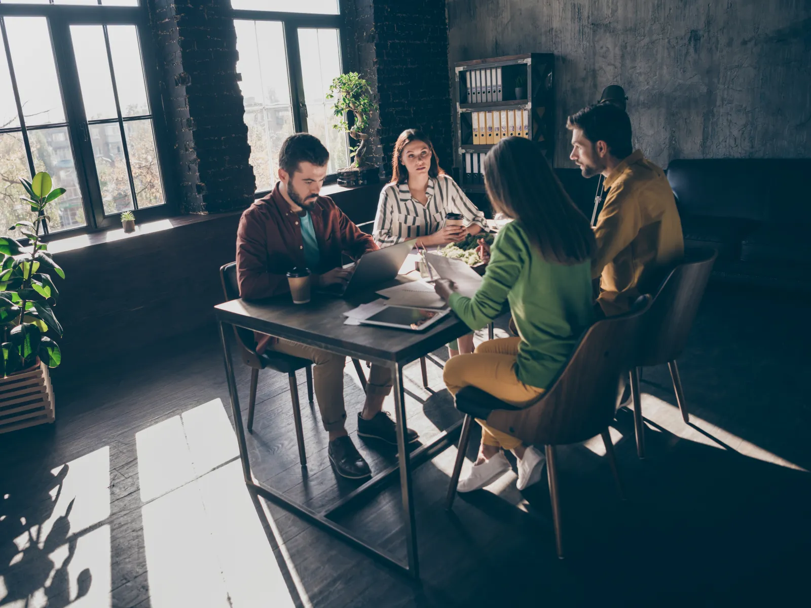 Four young professionals collaborating at a table in a modern office with large windows and natural light.