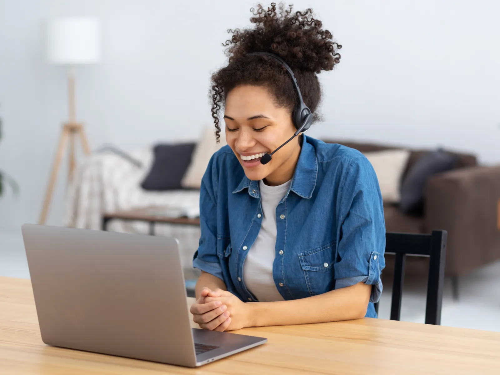 Smiling woman wearing headset video chatting on laptop at home, sitting at wooden table in cozy living room.
