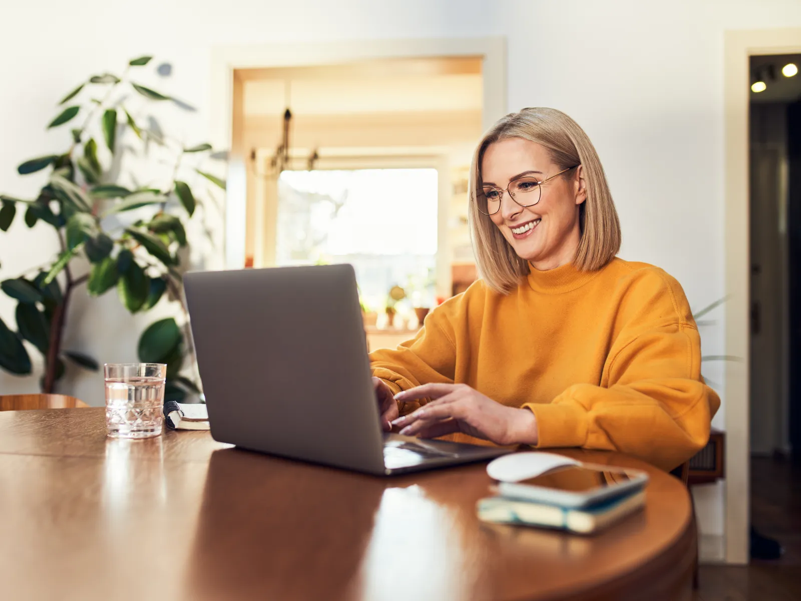 Smiling woman in glasses and orange sweater working on laptop at wooden table in cozy home office.