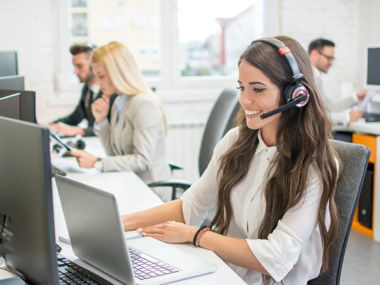 Smiling woman in headset working at a computer in a busy modern office with colleagues in the background.