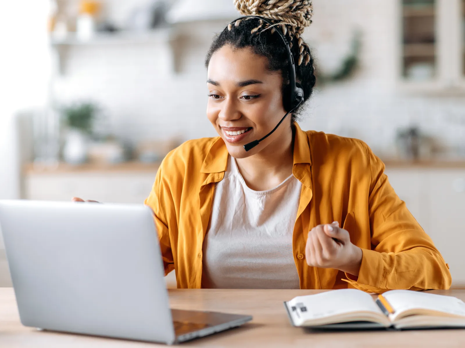 Smiling woman wearing headset using laptop and studying with open notebook at wooden table indoors.