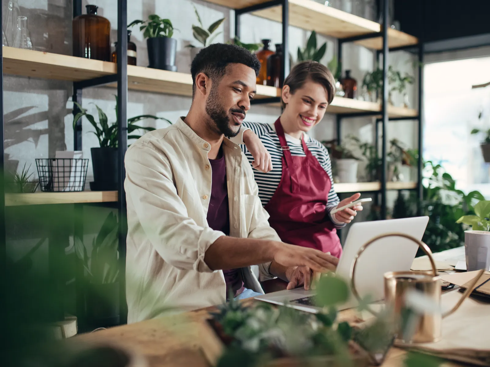 Man and woman working together on laptop in a plant-filled workspace with gardening tools and shelves behind them