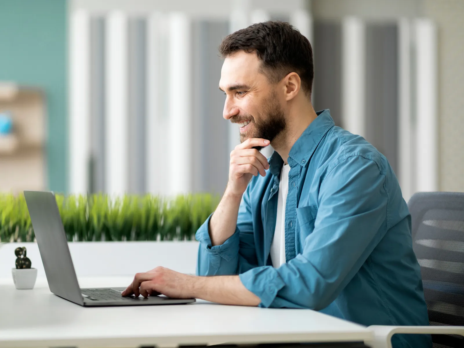 Man in blue shirt working on laptop at a clean white desk with green plants in a modern office space.