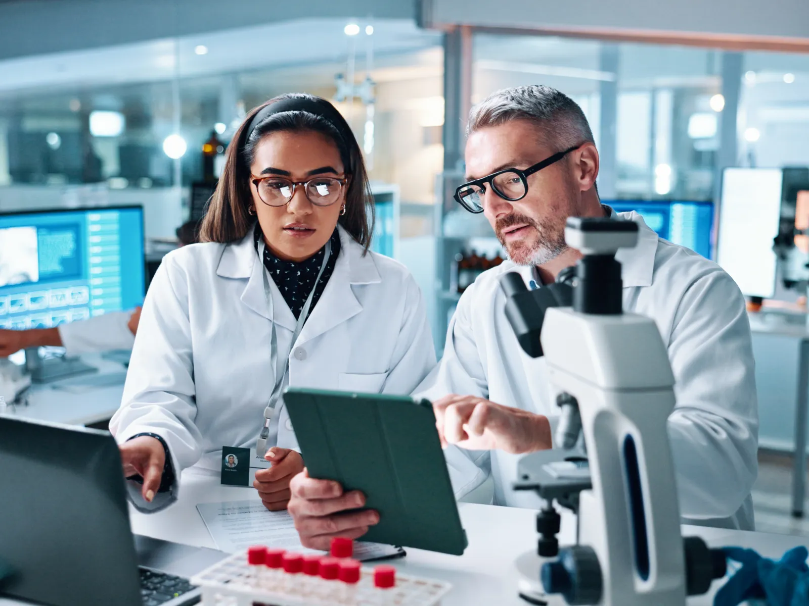 Two scientists in lab coats analyzing data on a tablet in a modern laboratory setting with microscope and equipment.