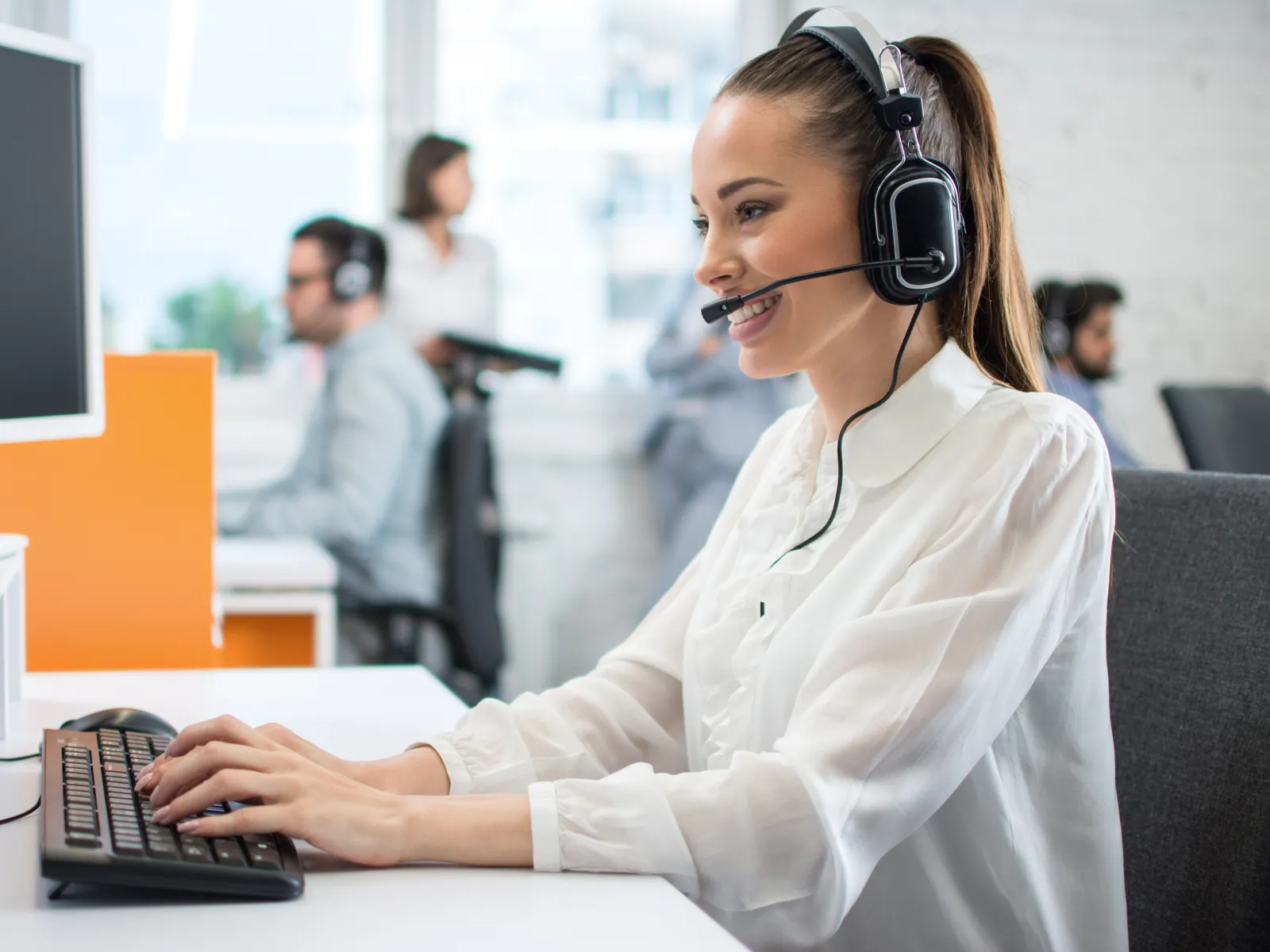 Smiling female customer service agent wearing headset typing on keyboard in busy modern office environment.