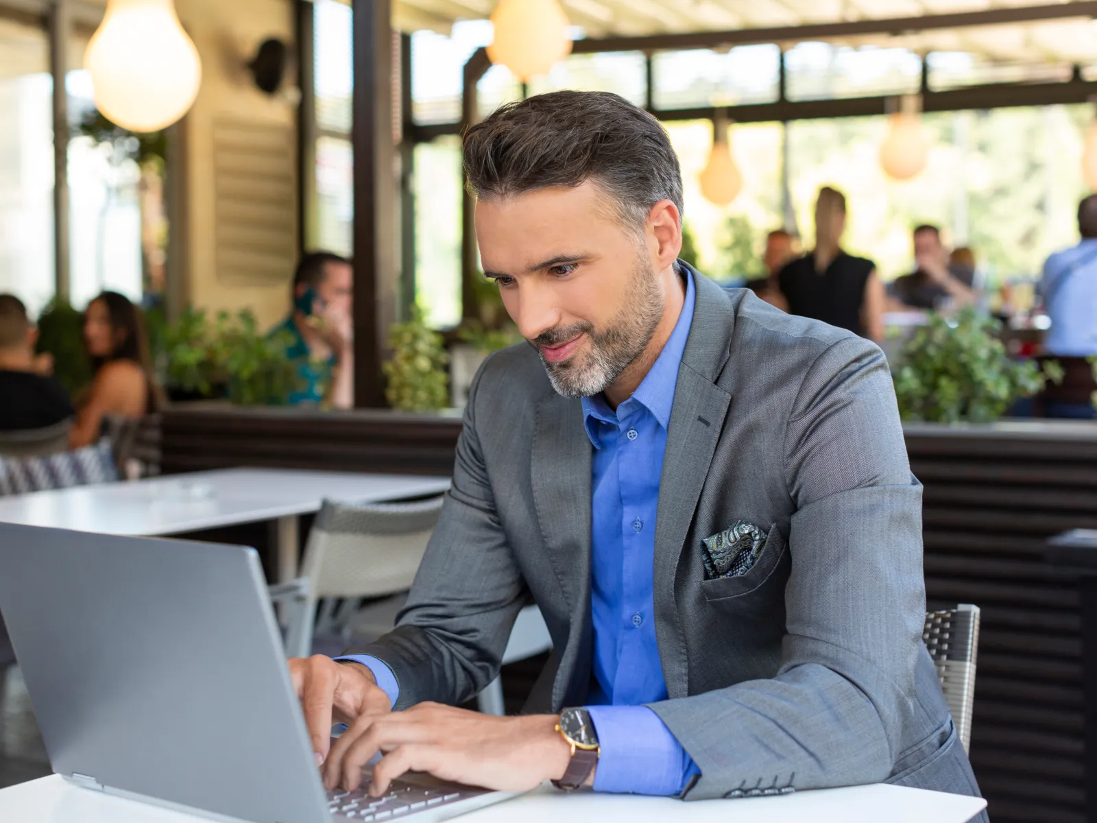 Businessman in a gray suit working on a laptop at an outdoor cafe with other people in the background