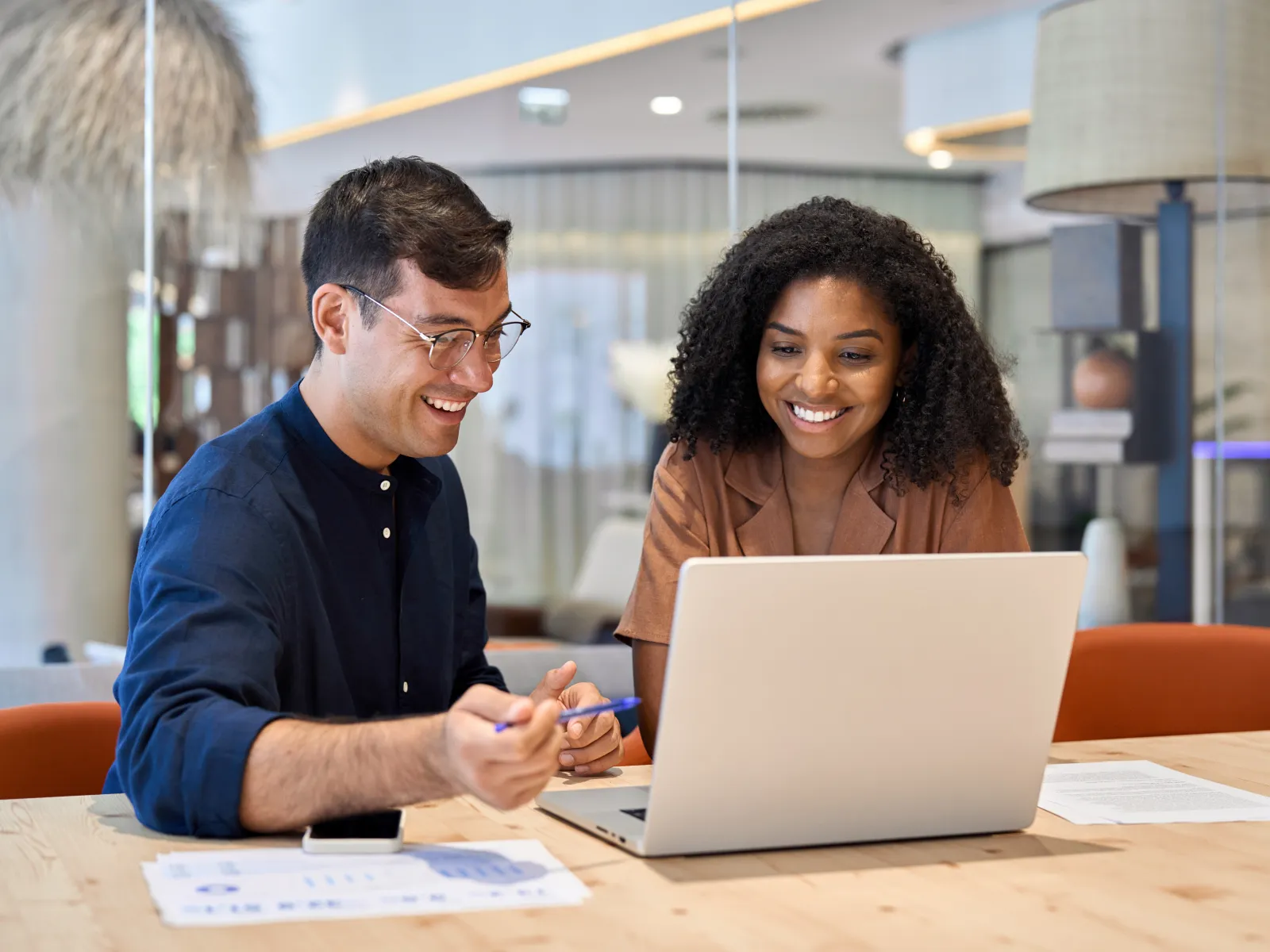 Two colleagues smiling and discussing work while looking at a laptop in a modern office setting