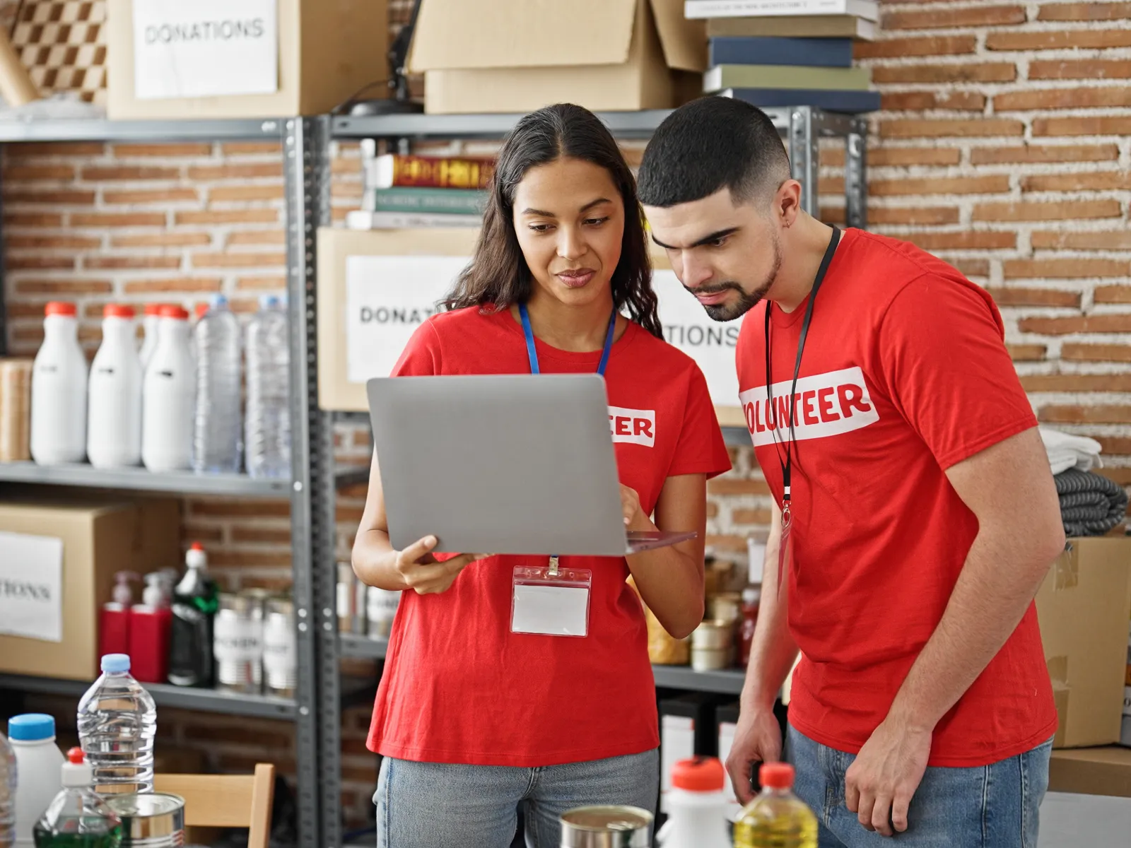Two volunteers in red shirts checking a laptop amid donation boxes and supplies in a storage room.