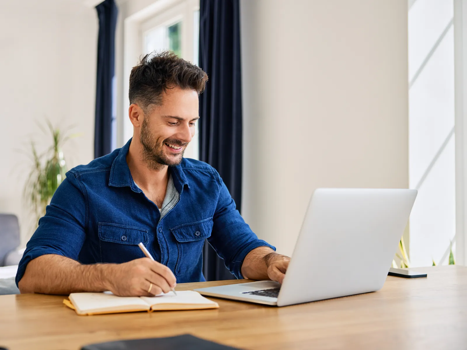 Man in blue shirt working on laptop and taking notes at wooden desk in bright room with plants.