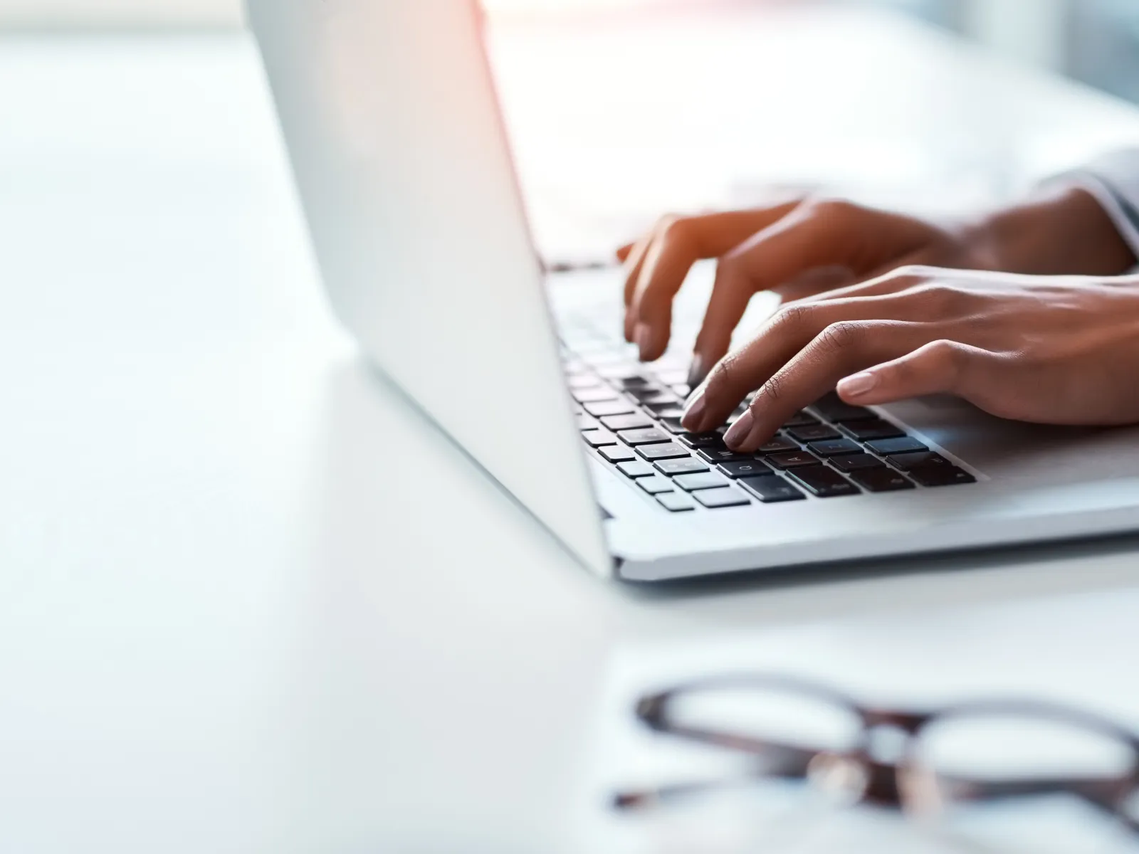 Hands typing on a laptop keyboard with glasses on a white desk in a bright workspace.