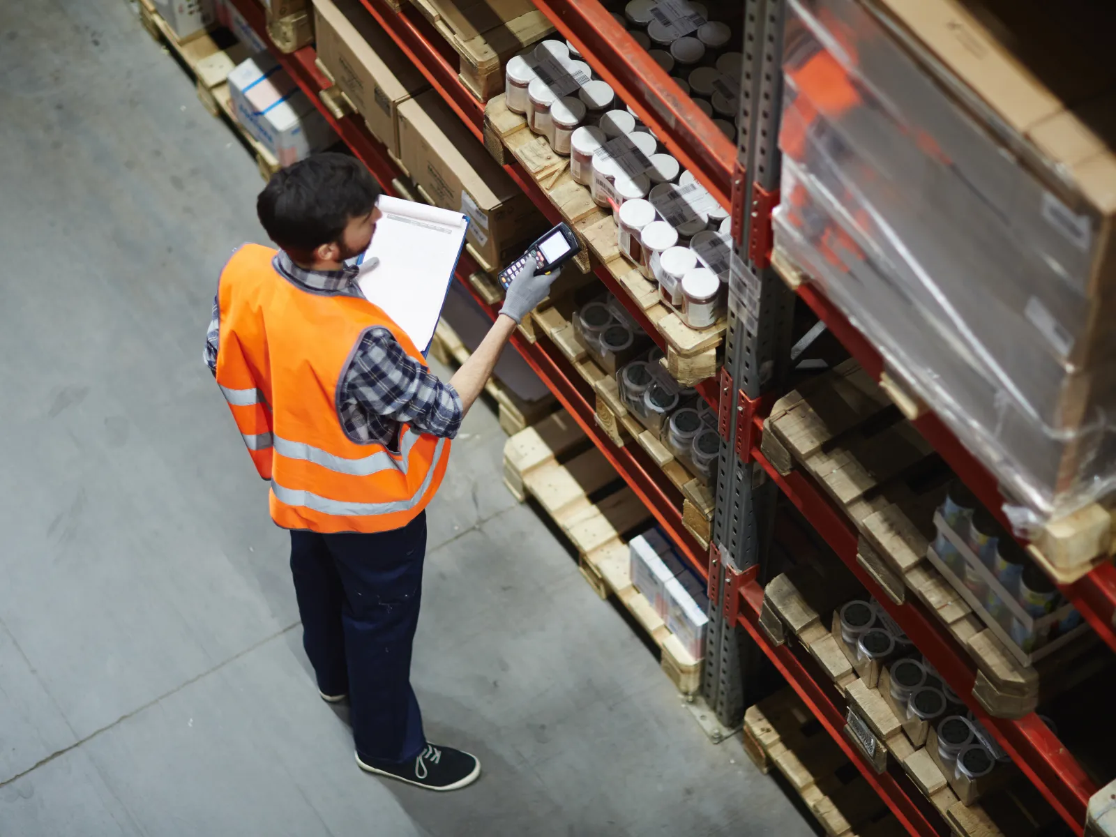 Warehouse worker in safety vest scanning inventory items on shelves with handheld device and clipboard