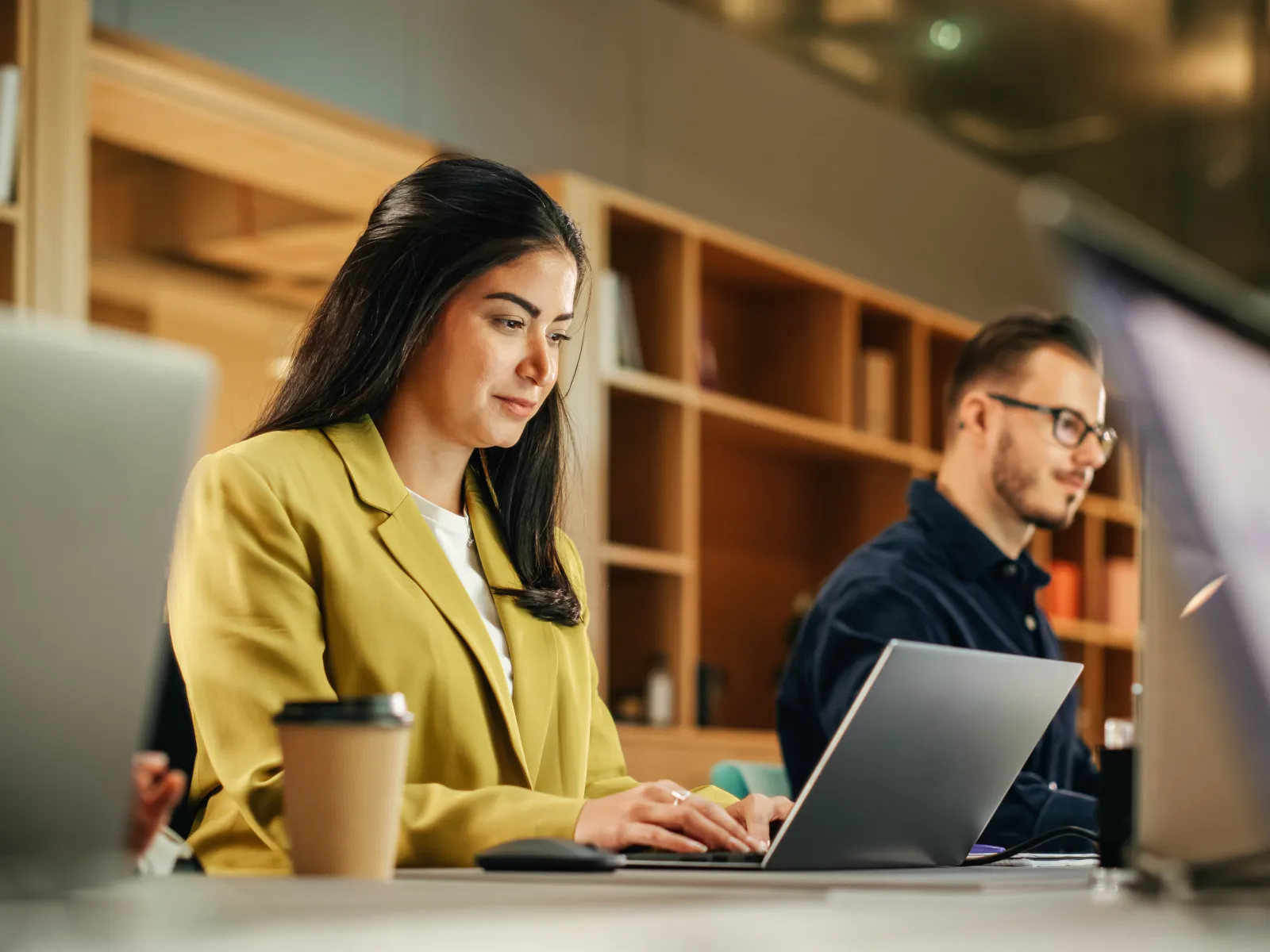 Young professionals focused on laptops working together in a modern office with wooden shelves and coffee cups.