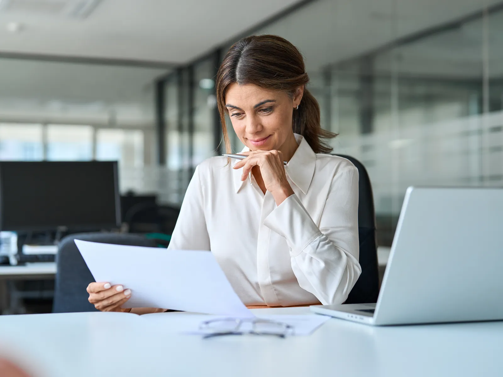 Businesswoman in white shirt reviewing documents thoughtfully at office desk with laptop and eyeglasses.
