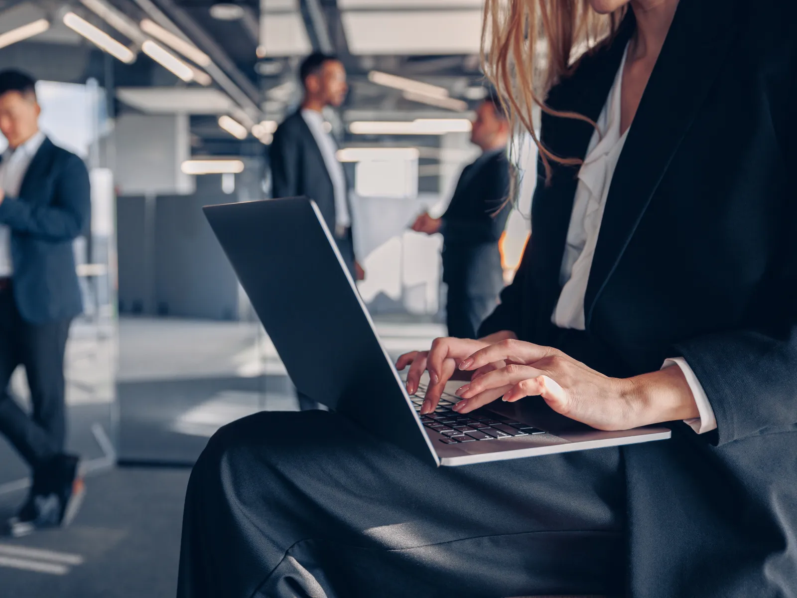 Businesswoman using laptop in modern office while colleagues talk in background during workday