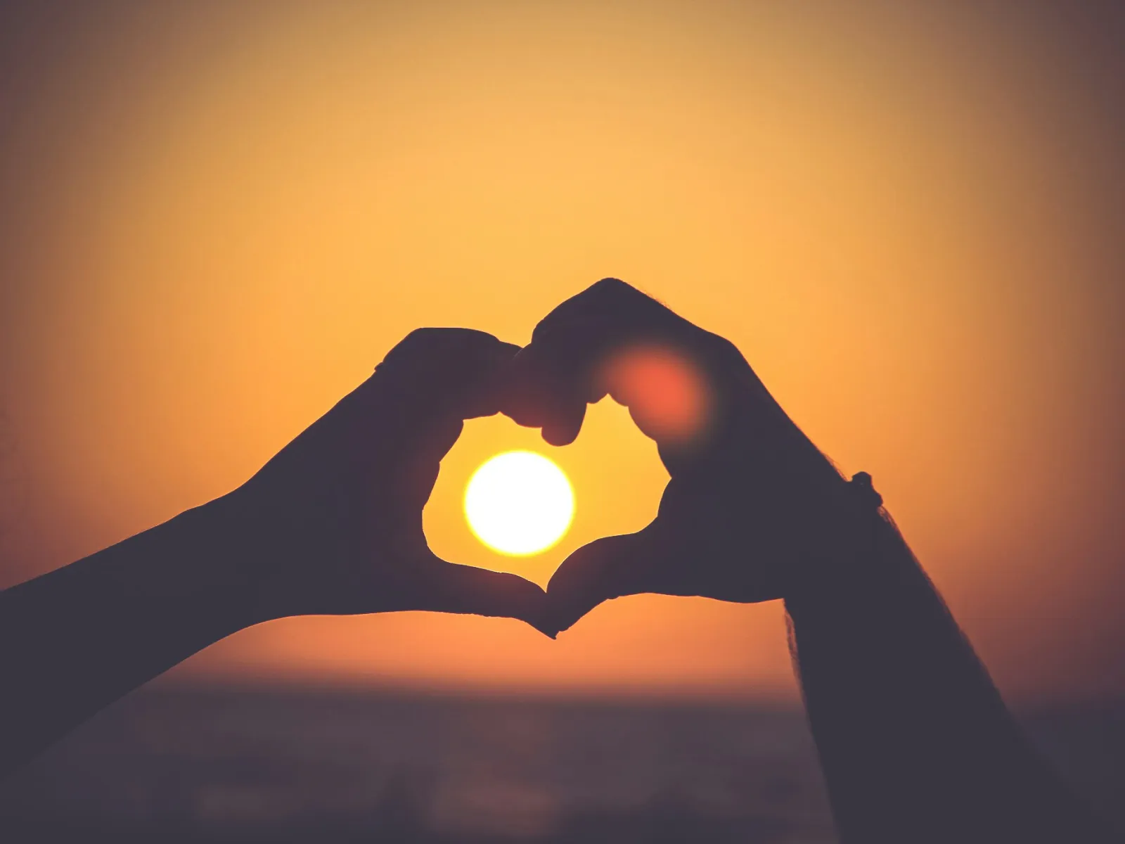 Silhouetted hands forming a heart shape around a sunset over the ocean with warm orange sky.