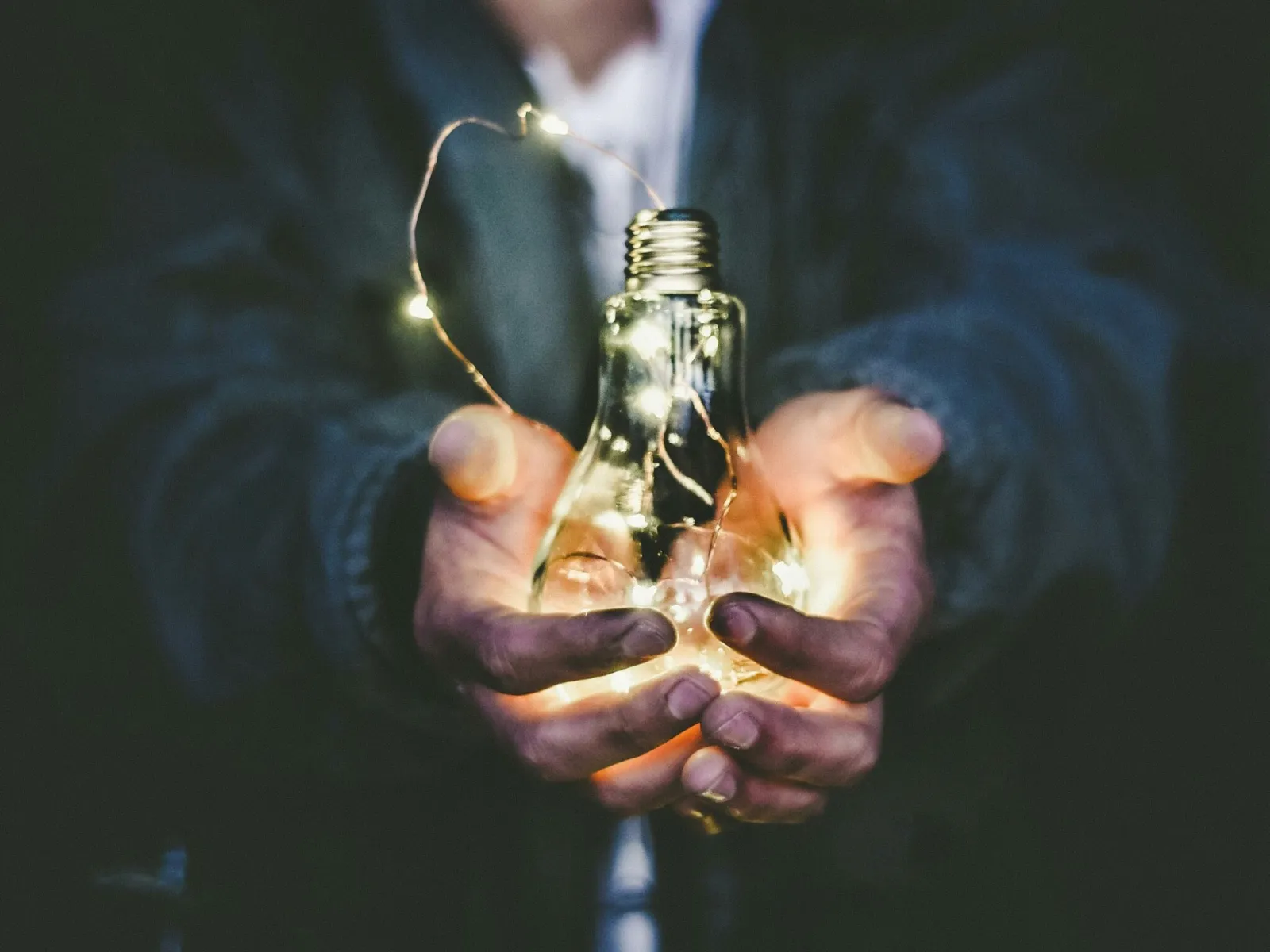 Person holding a glowing light bulb filled with string lights against a dark background.