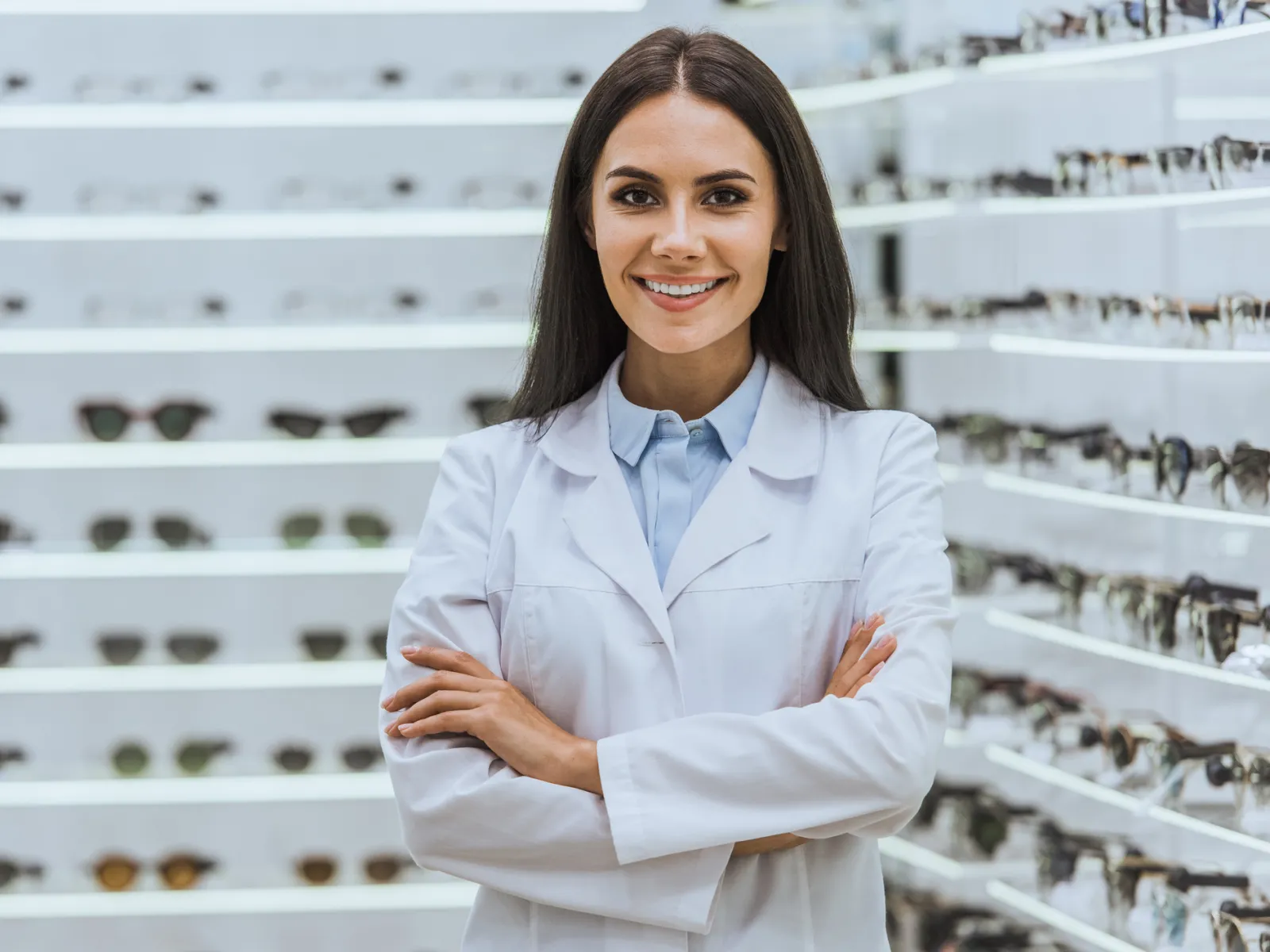 A smiling optometrist in a lab coat stands confidently in front of a display of sunglasses.